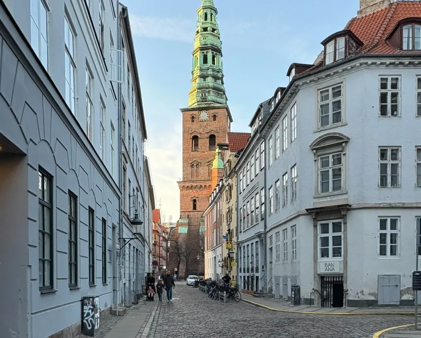 Mennesker vandrer langs en middelalderbygning, der er i flere etager og har et klokketårn, står i et ældre byområde med brostensbelagte gader