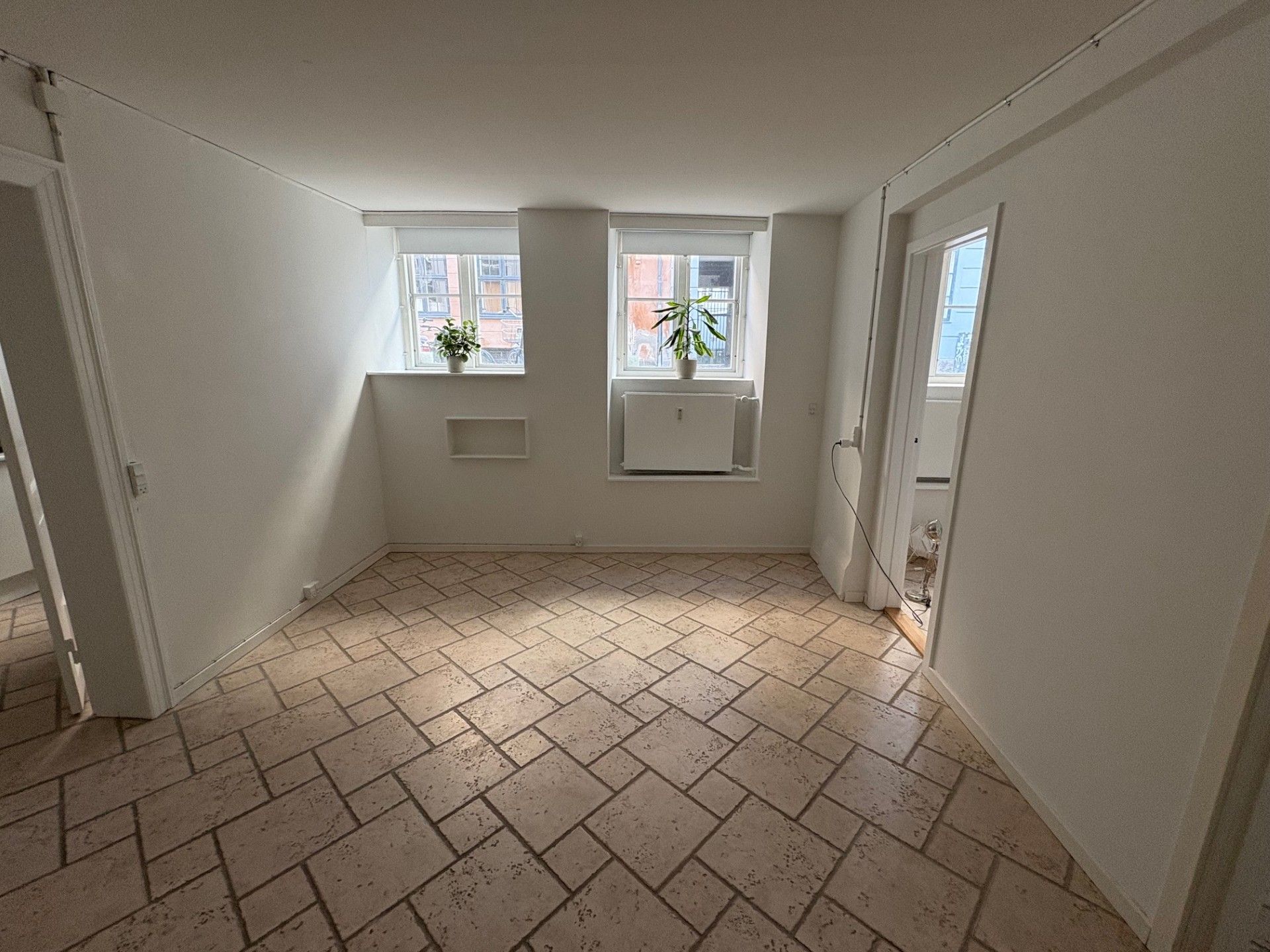 Et tomt kontorlokale med klinkergulv og hvide vægge. To vinduer med plantepotter og radiatorer under dem. Døre på hver side og en åben dør i midten.