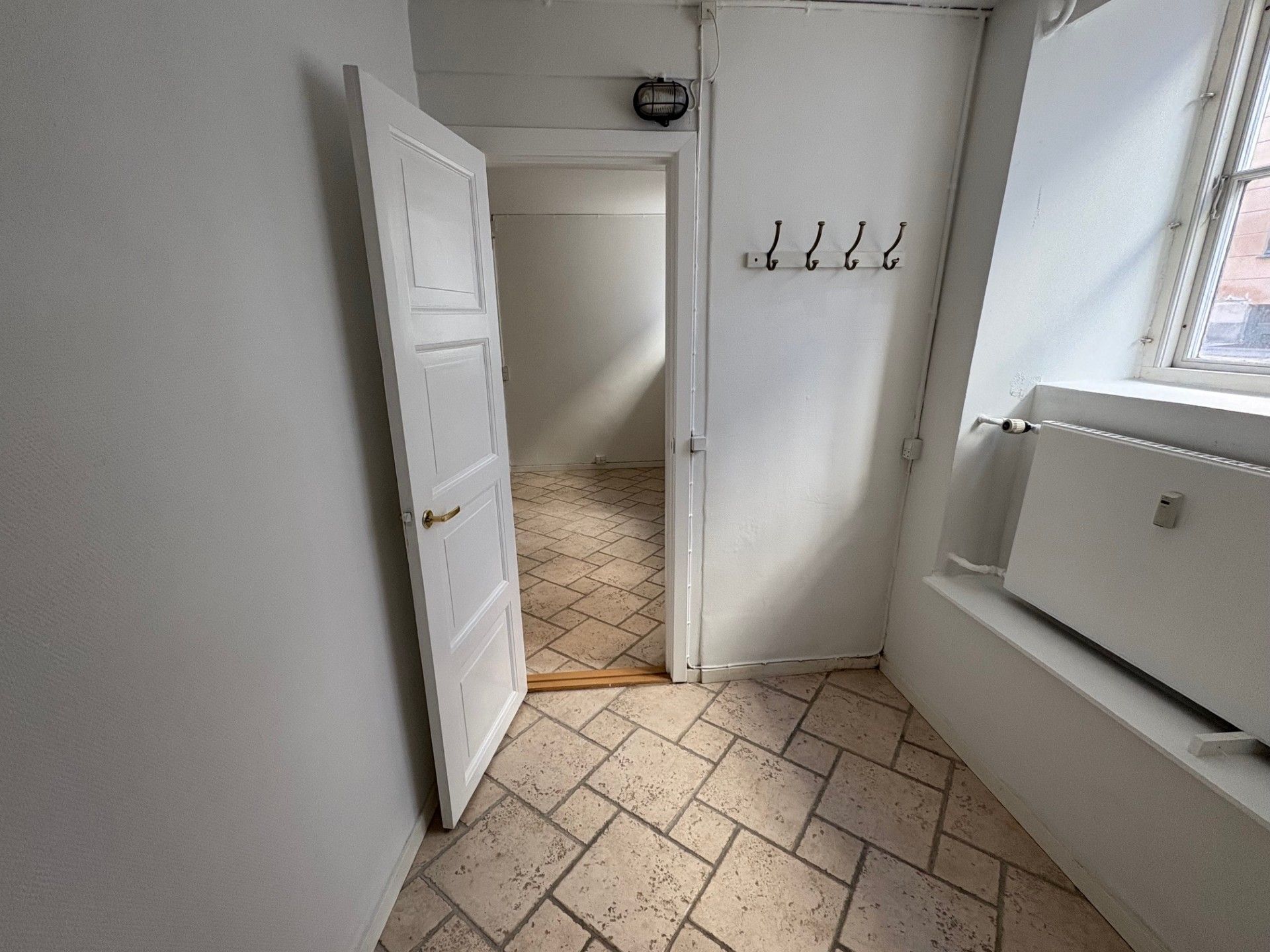 Hvidt vægmaleri med åben dør og lyse gulvfliser, tre hængeklaer og vinduesruder med udsigt til byen og trægulv i gangen