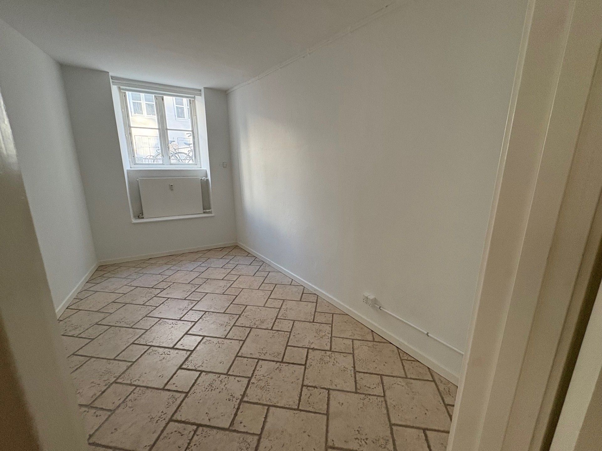 Lejlighed med gult belagt gulv og hvidmalede vægge samt vindue med metalruder og gardiner. Loftet er hvidmalet og der er radiator under vinduet.