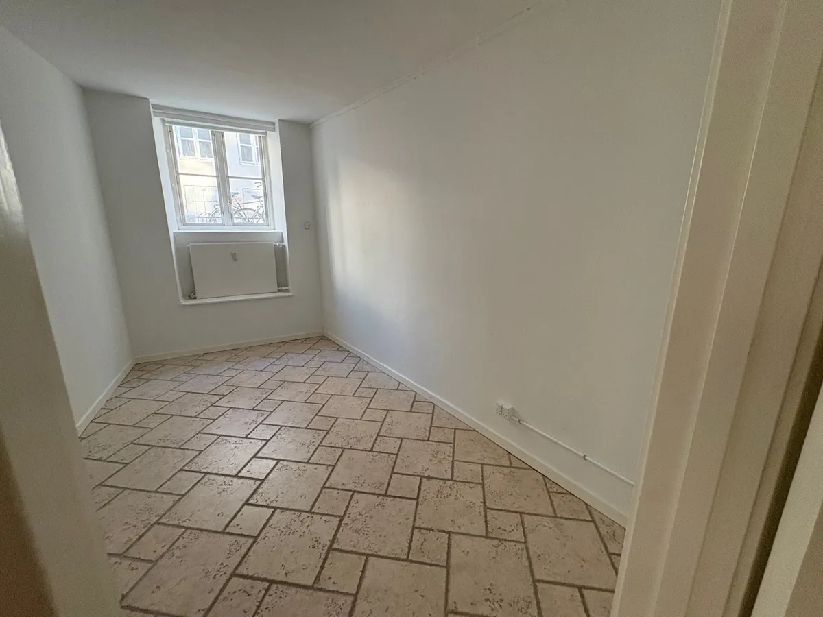 Lejlighed med gult belagt gulv og hvidmalede vægge samt vindue med metalruder og gardiner. Loftet er hvidmalet og der er radiator under vinduet.