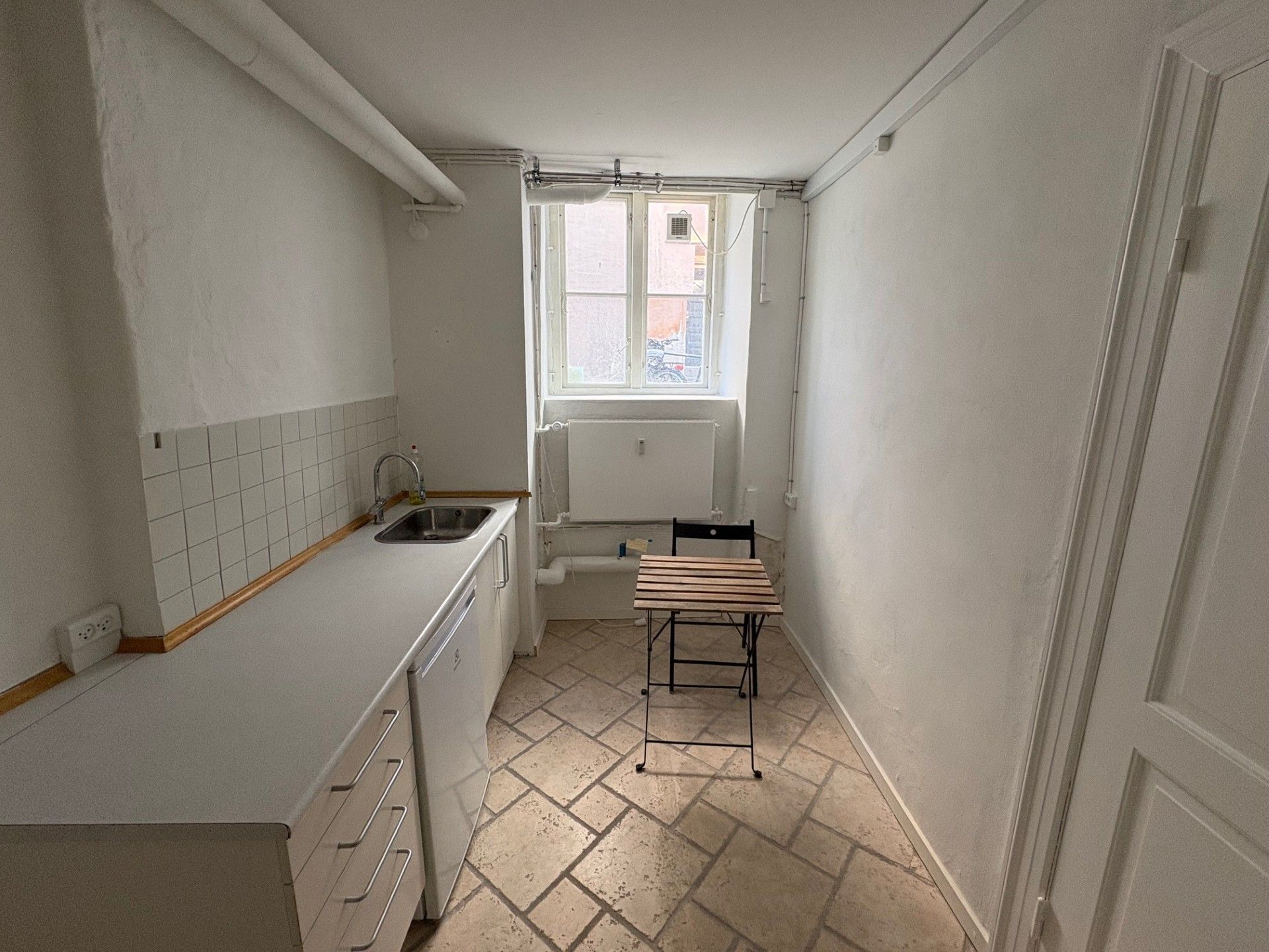 En lille køkken med hvidt loft, hvide vægge, lysegrå gulvfliser og lysegråt bord med stole uden for