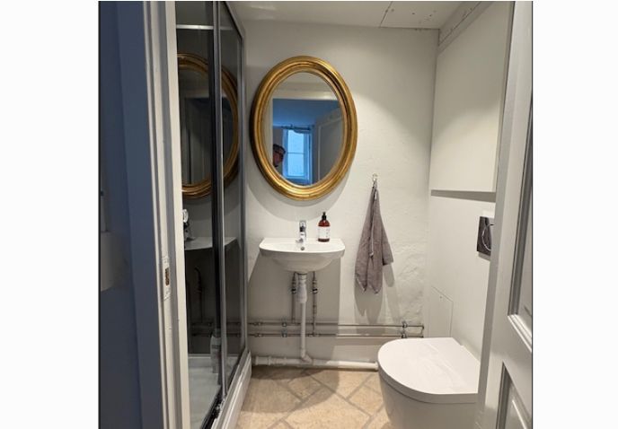 En lille hvid badeværelse med et rundt guldfarvet spejl, hvidt toilet, vask og en hængende grå håndklæde