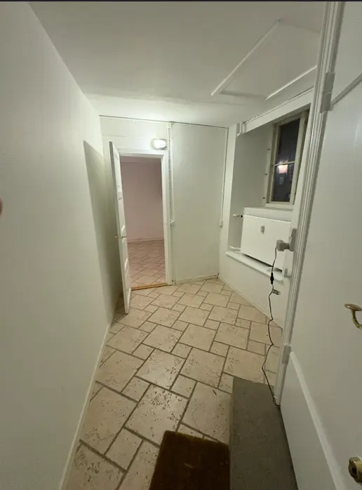 Et rummeligt kontor med to åbne døre og en indvendig vindueskilt. Lys er tændt inde, og der er et gulvtæppe ved en dør. Væggene er hvide, og der er en hvid radiator under vinduet.