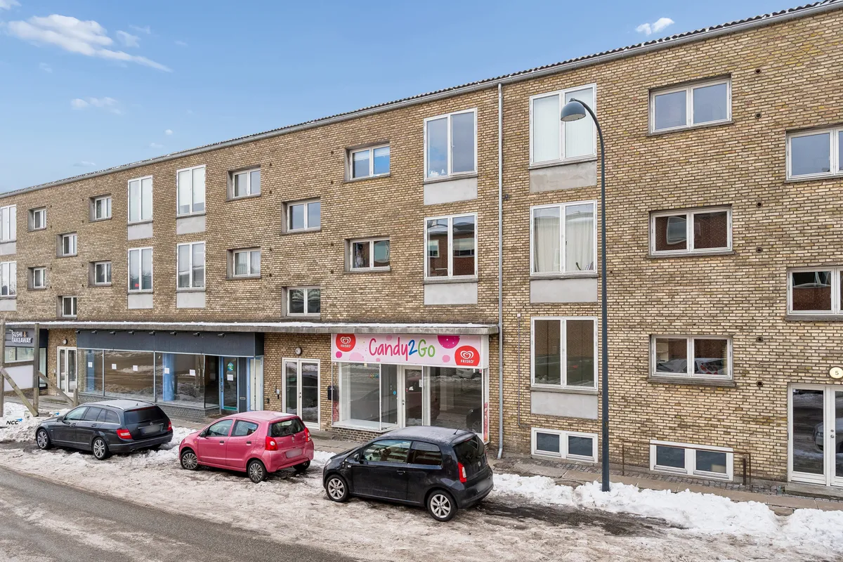 Butik med Candy2Go-skilt, og tre biler parkeret foran på Snogegårdsvej. Vintervejr med sne og skyer på himlen.