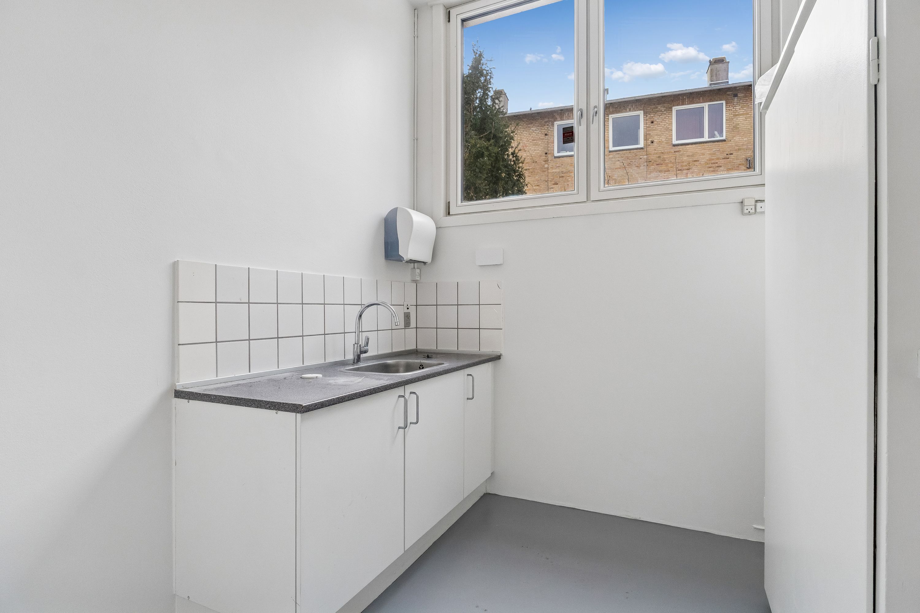 En lille tømmerkøkken med hvide vægge, en sink, en håndvask og en håndklædeholder. Vinduet ud mod en bygning med røde mursten og tre vinduer.