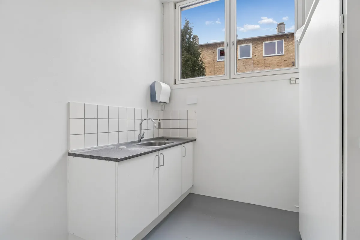 En lille tømmerkøkken med hvide vægge, en sink, en håndvask og en håndklædeholder. Vinduet ud mod en bygning med røde mursten og tre vinduer.