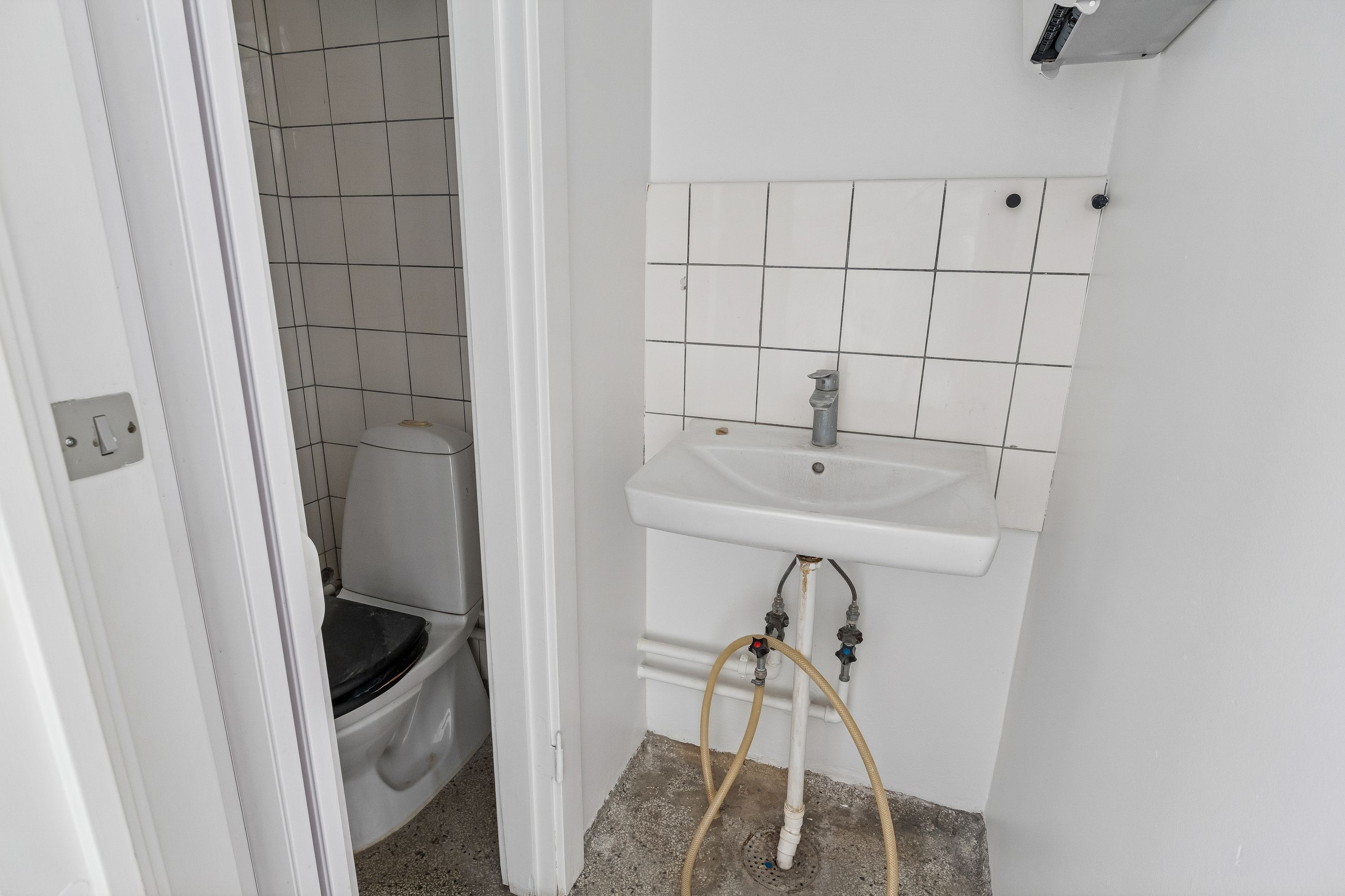 Hjørne af et lille badeværelse med hvidt toilet og hvidt vask, samt en gul slange forbundet til vandforsyningen