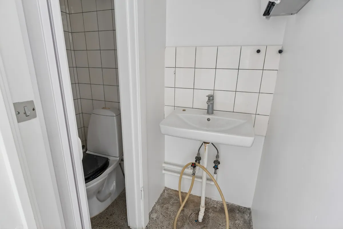 Hjørne af et lille badeværelse med hvidt toilet og hvidt vask, samt en gul slange forbundet til vandforsyningen