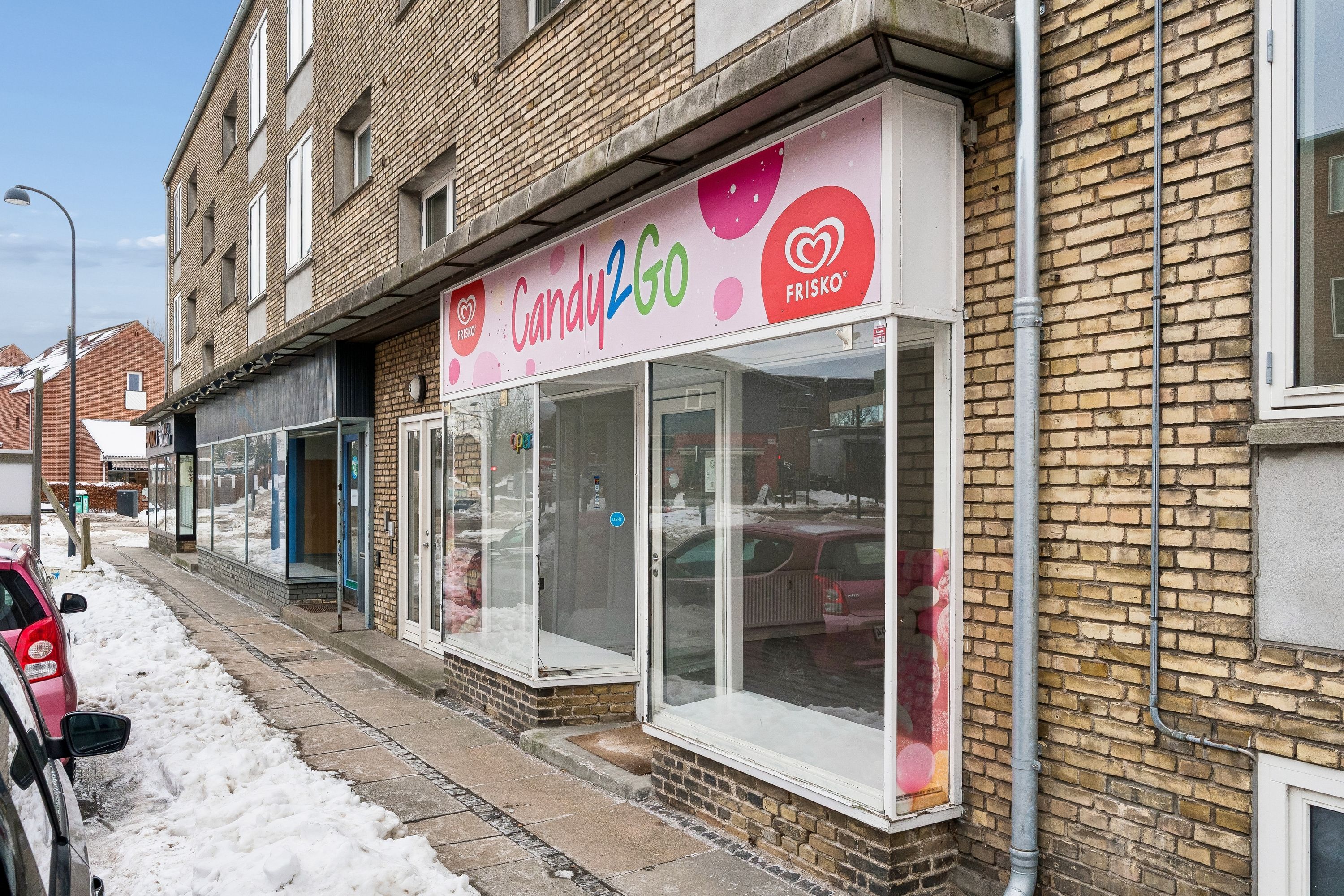 En Candy2Go-butik med glasruder og en lyserød facade, som er bygget i røde mursten. Butikken er placeret på en snemørk vej med parkerede biler på siderne.