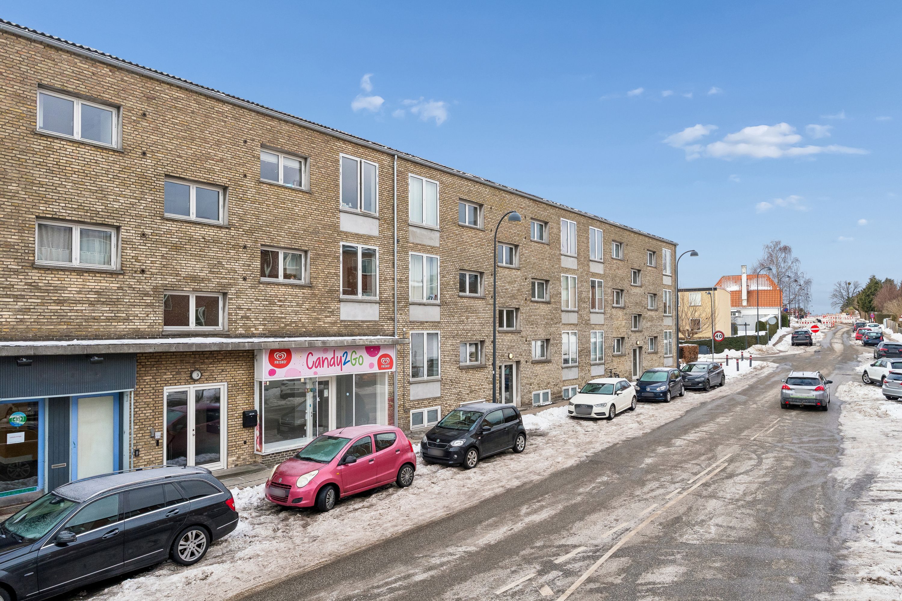 Et stort rødt murstenshus med mange vinduer og udsalgslokaler, hvor der er parkeret biler på begge sider af vejen i et kold vinterdag