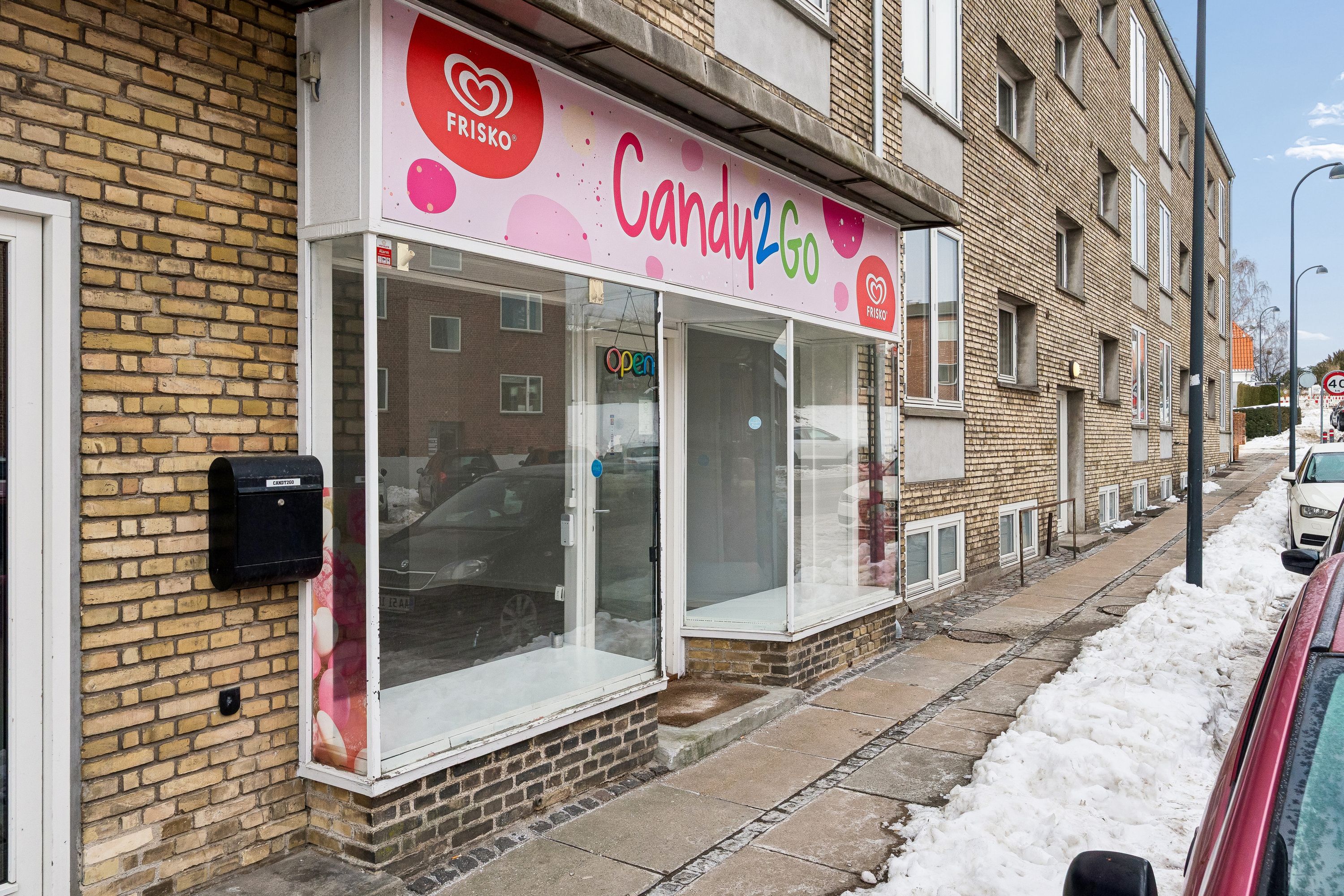 Candy2Go butik med pæn glasfacade og åben dør på Snogegårdsvej. Butikken har en 40 km/t skilt, og der er biler parkeret på hjørnet.