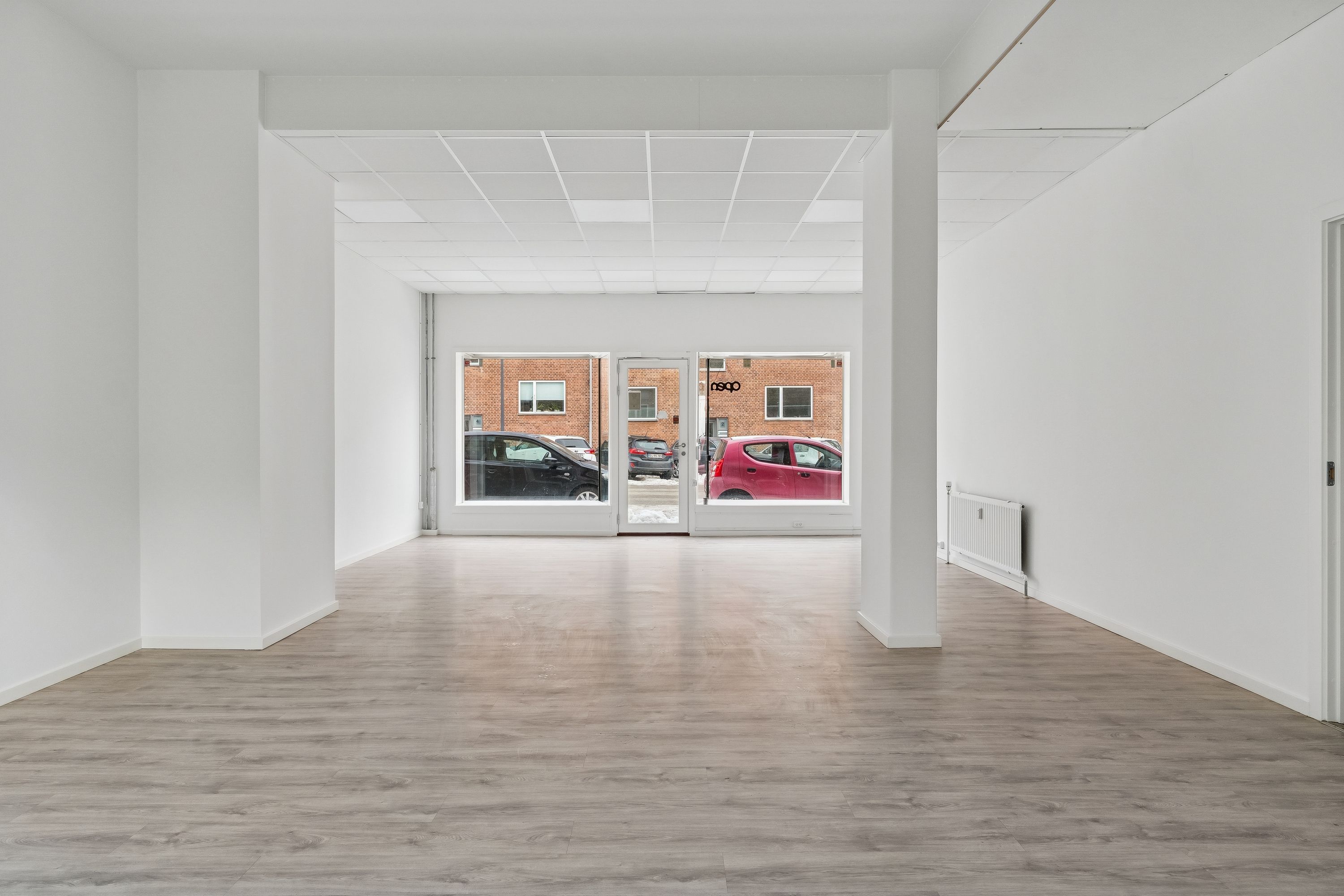 Et stort og lyst lokale med gulvbelægning og hvide vægge. Der er et glasdørsystem og vinduer med udsigt til en parkeringsplads. Et hvidt radiator står i hjørnet