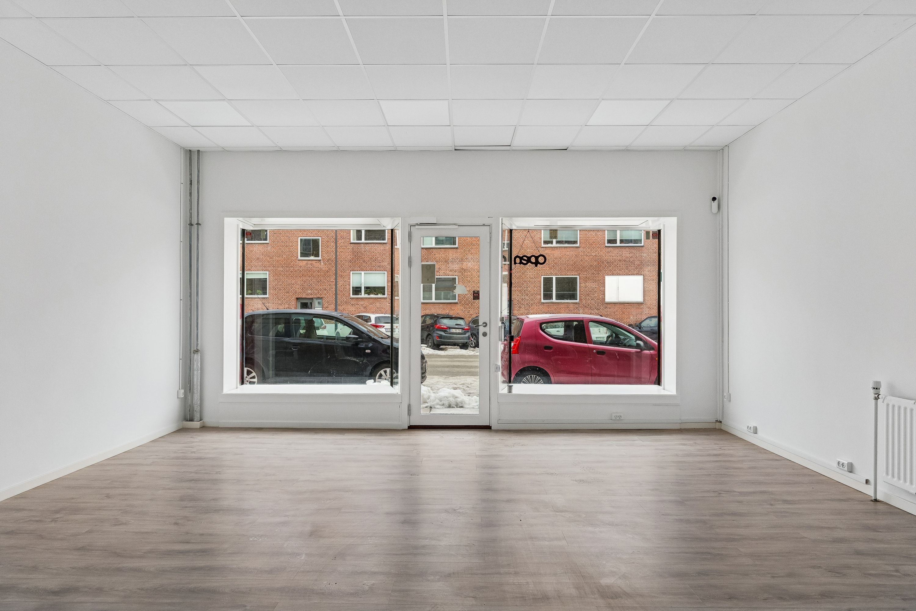 En stor butik med to store vinduer og en glasdør, der vender ud mod en parkeringsplads med biler og en rødmalet bil. Væggene og loftet er hvide, og der er en radiator ved siden af døren