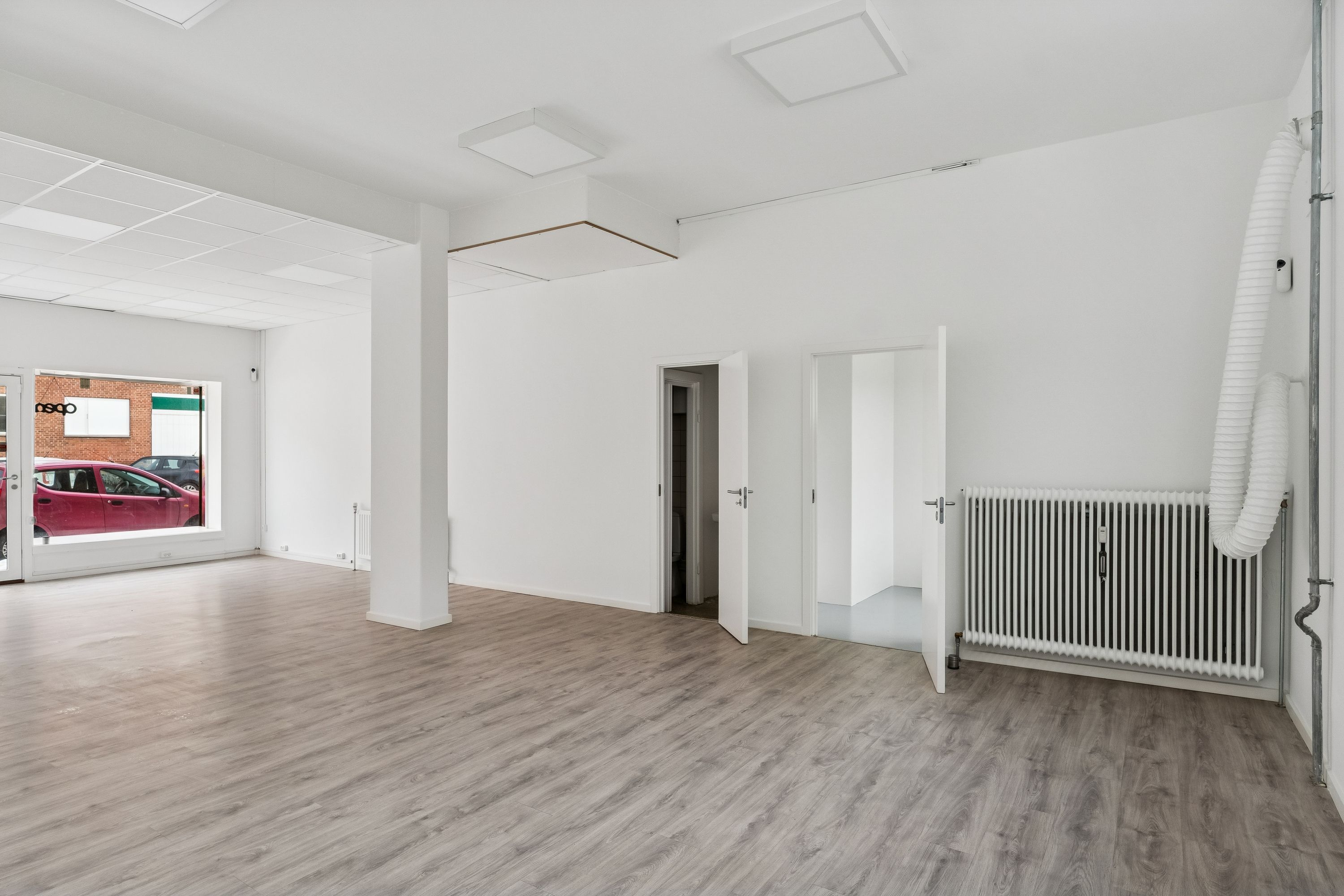 Store, lyse lokaler med højt til loftet og lyse gulvbelægning. Her er også radiatorer og en åben dør til et mindre rum. Der er en bilparkeret udenfor.