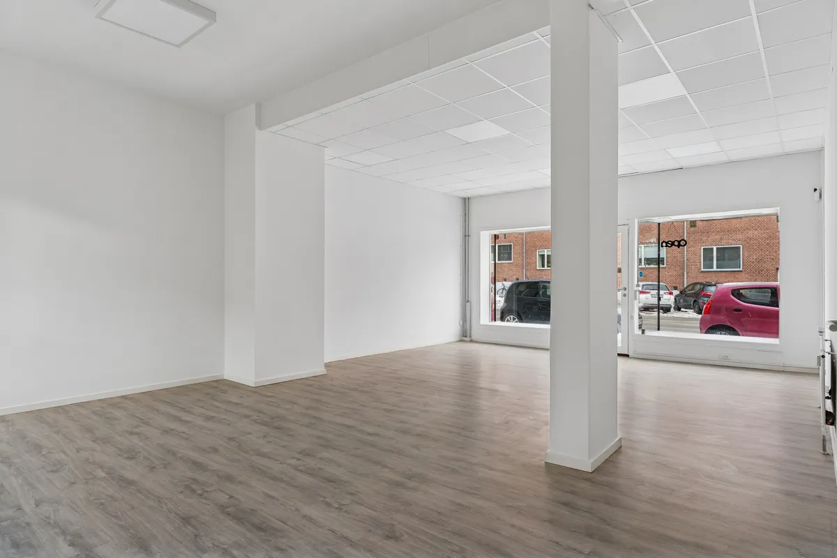 Butik med hvide vægge, trægulv og store vinduer, der vender ud mod en parkeringsplads med biler. Pladsen er tom, og der er ingen møbler.