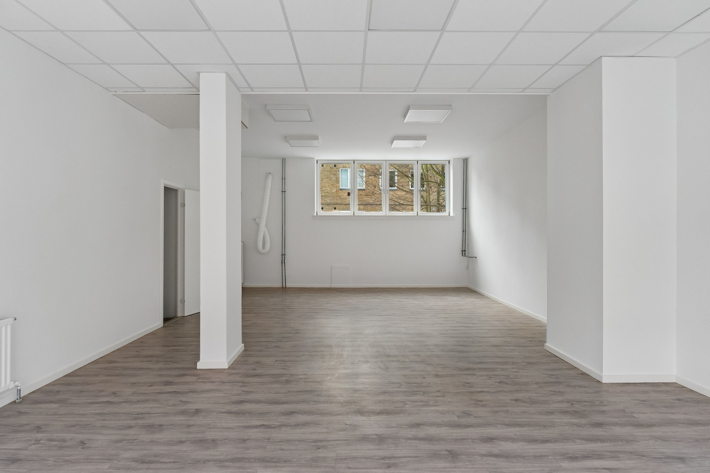 Ledig butikslokale med 81 m², hvid væg og lysegrå gulvbelægning, samt en rektangulær vinduesfacade med glas. Rummet er forsynet med hvide lofter, hvilket giver en minimalistisk udformning.