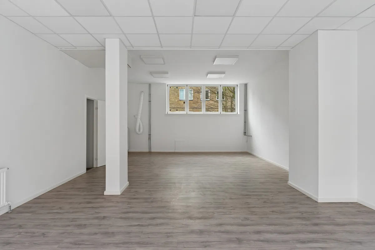 Ledig butikslokale med 81 m², hvid væg og lysegrå gulvbelægning, samt en rektangulær vinduesfacade med glas. Rummet er forsynet med hvide lofter, hvilket giver en minimalistisk udformning.