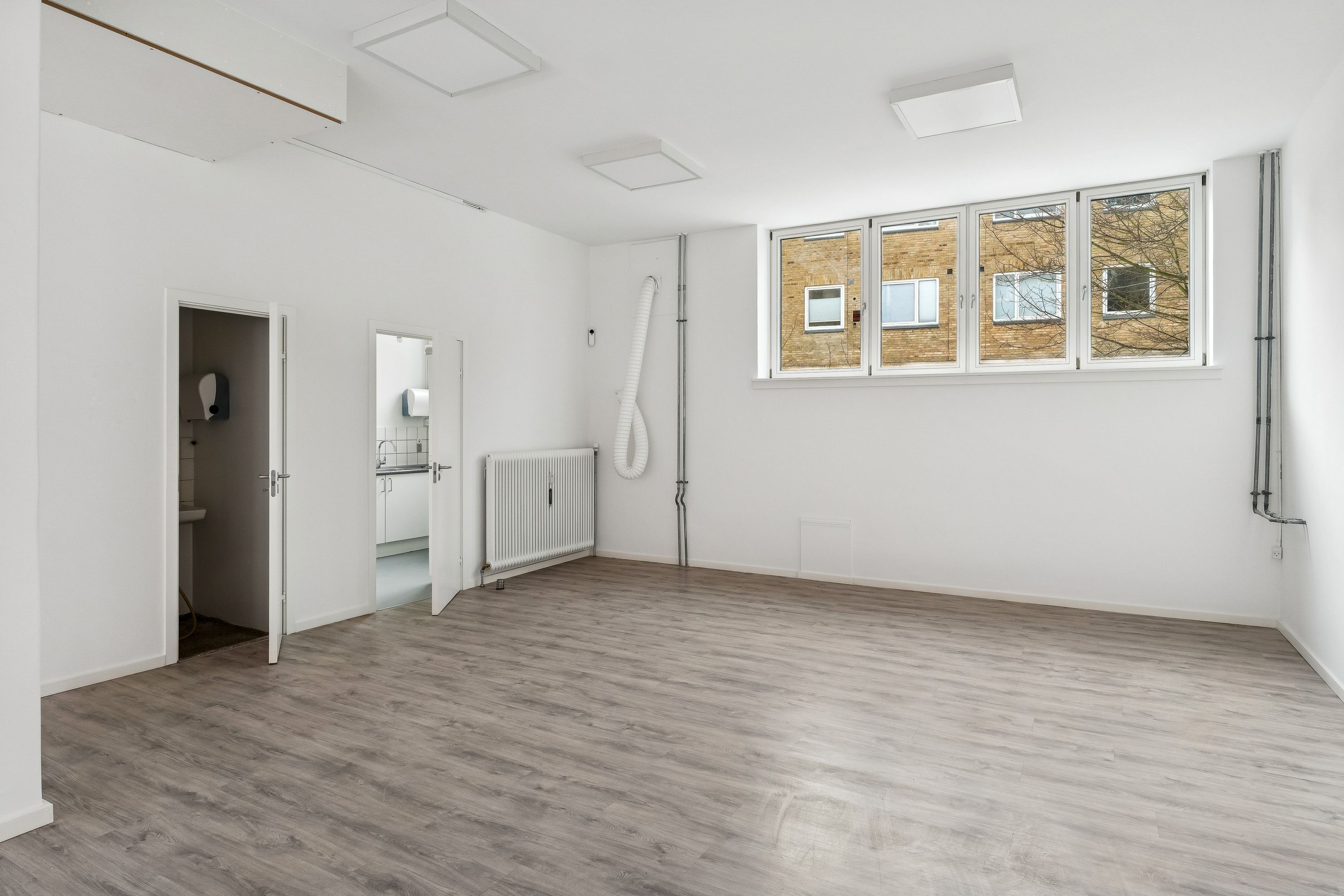 Lejlighed med åben dør, hvid væg og loft og lyse gulvbelægning. Ventilationsrør og radiator er placeret mod væggen. Et vindue med glasfacade og udsigt til en bygning med røde mursten.