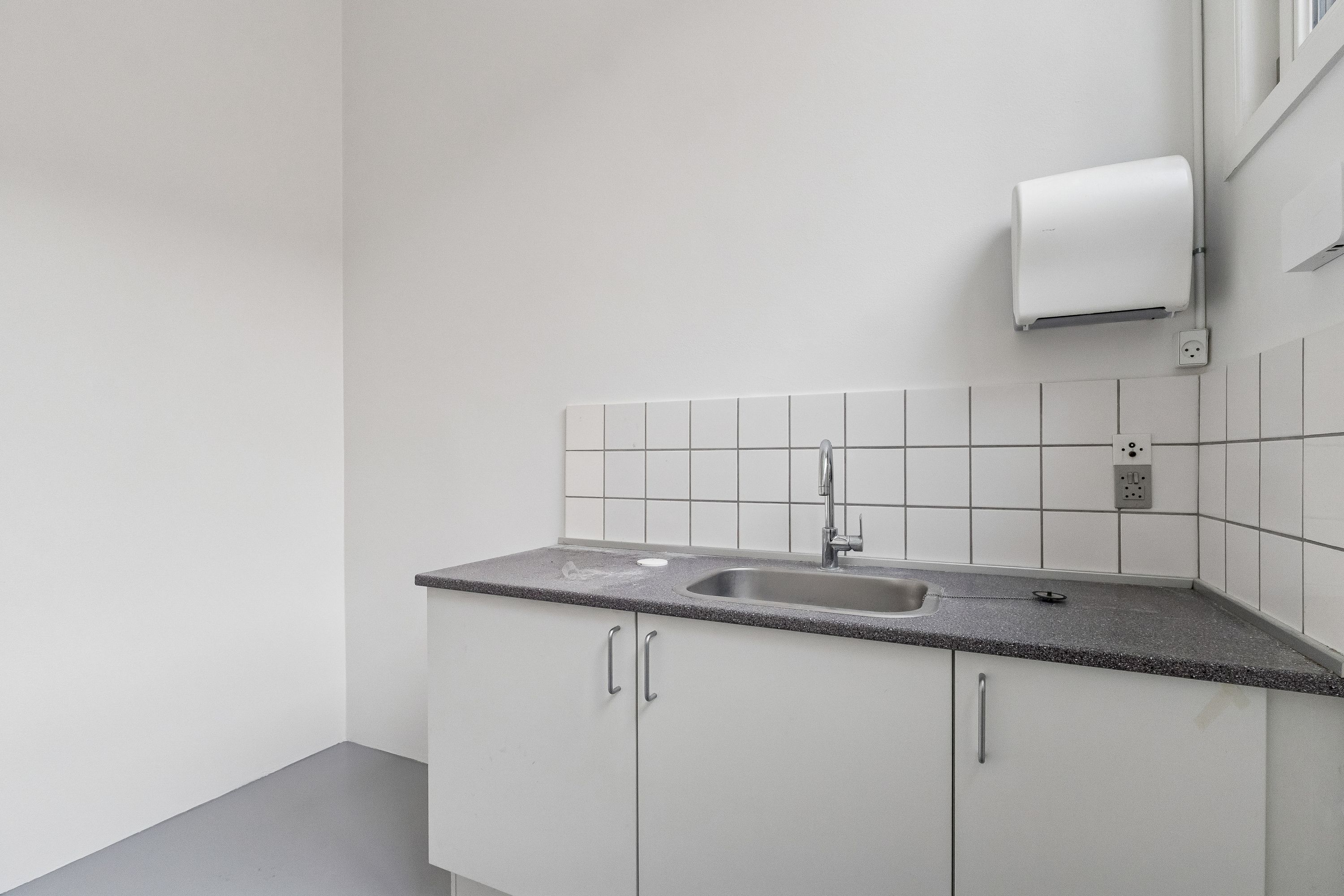 En lille, hvid og ren forretningskøkken med hvide vægge og loft, et sædvanligt håndvask og håndklædeholder, samt elektriske stik og en vinduesåbningsmekanisme