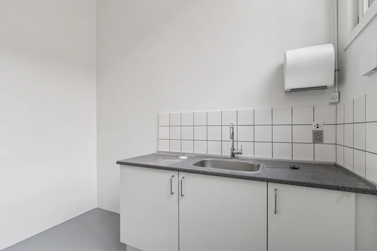 En lille, hvid og ren forretningskøkken med hvide vægge og loft, et sædvanligt håndvask og håndklædeholder, samt elektriske stik og en vinduesåbningsmekanisme