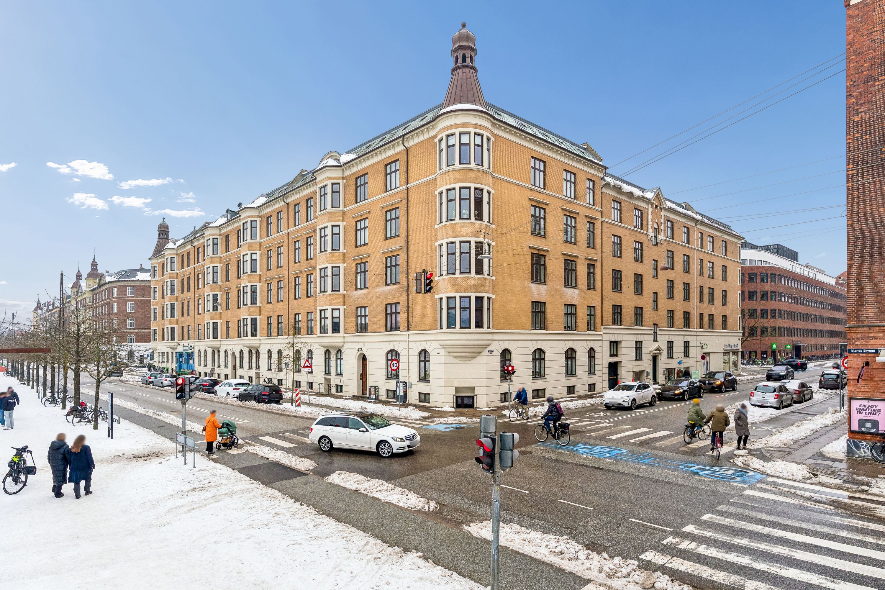 Køre på Islands Brygge med bil og cykel, og fodgængere går på sne. En stor bygning med flere etaper og hjørne med skrånende tag. Køretøjer parkeret langs gaden.