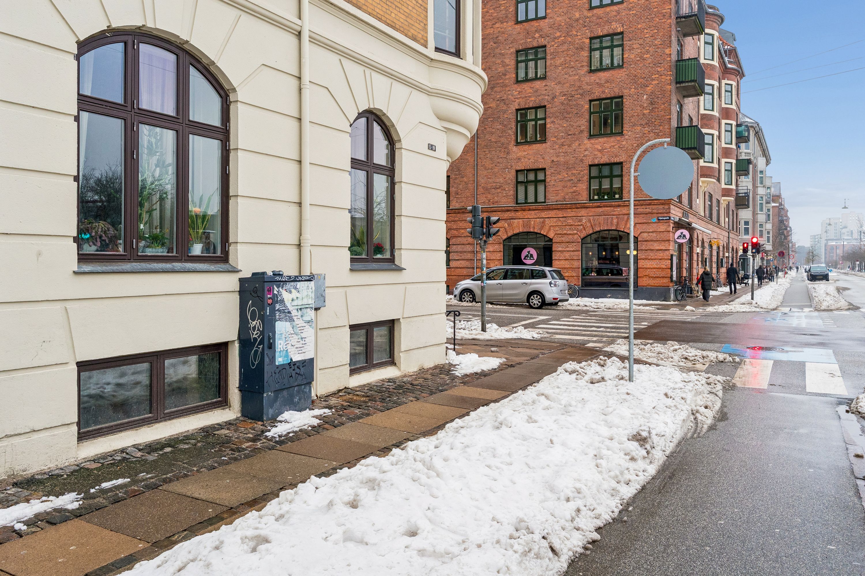 Gade med sne og mennesker, skovejern, trafiklys, biler og bygninger med vinduer og balkoner i baggrunden