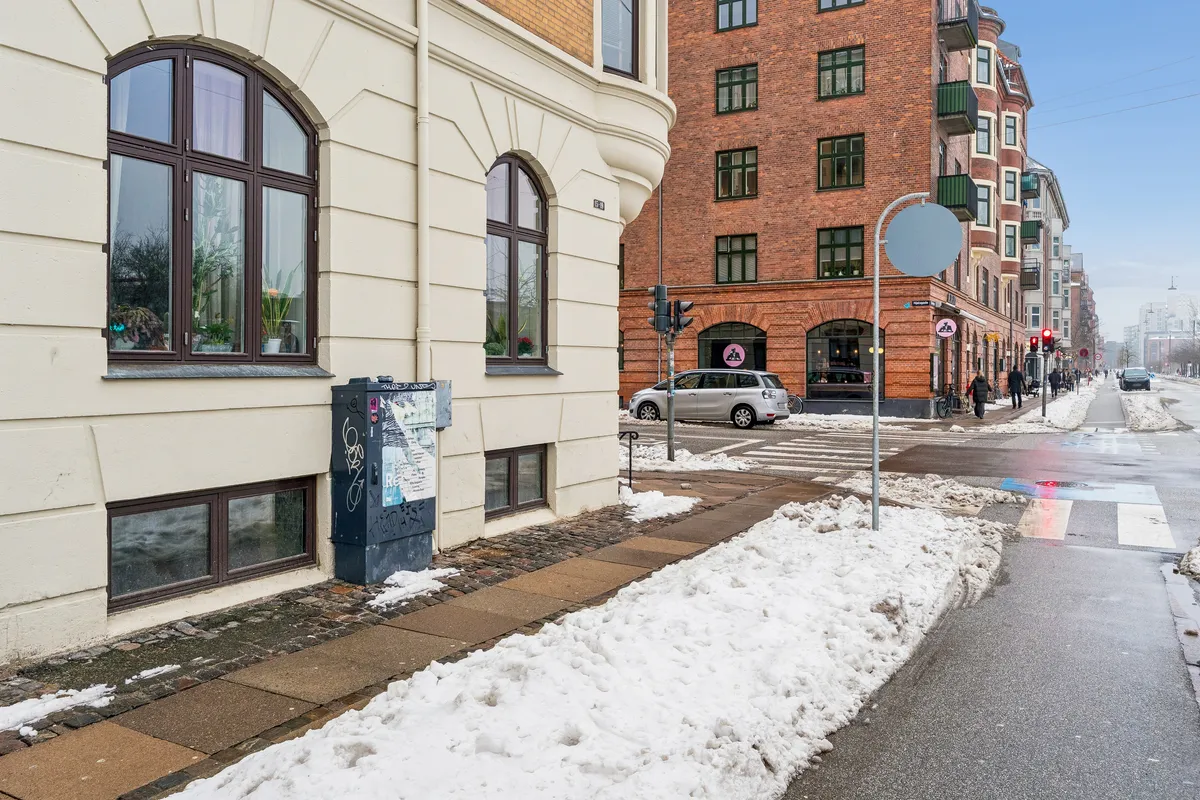 Gade med sne og mennesker, skovejern, trafiklys, biler og bygninger med vinduer og balkoner i baggrunden