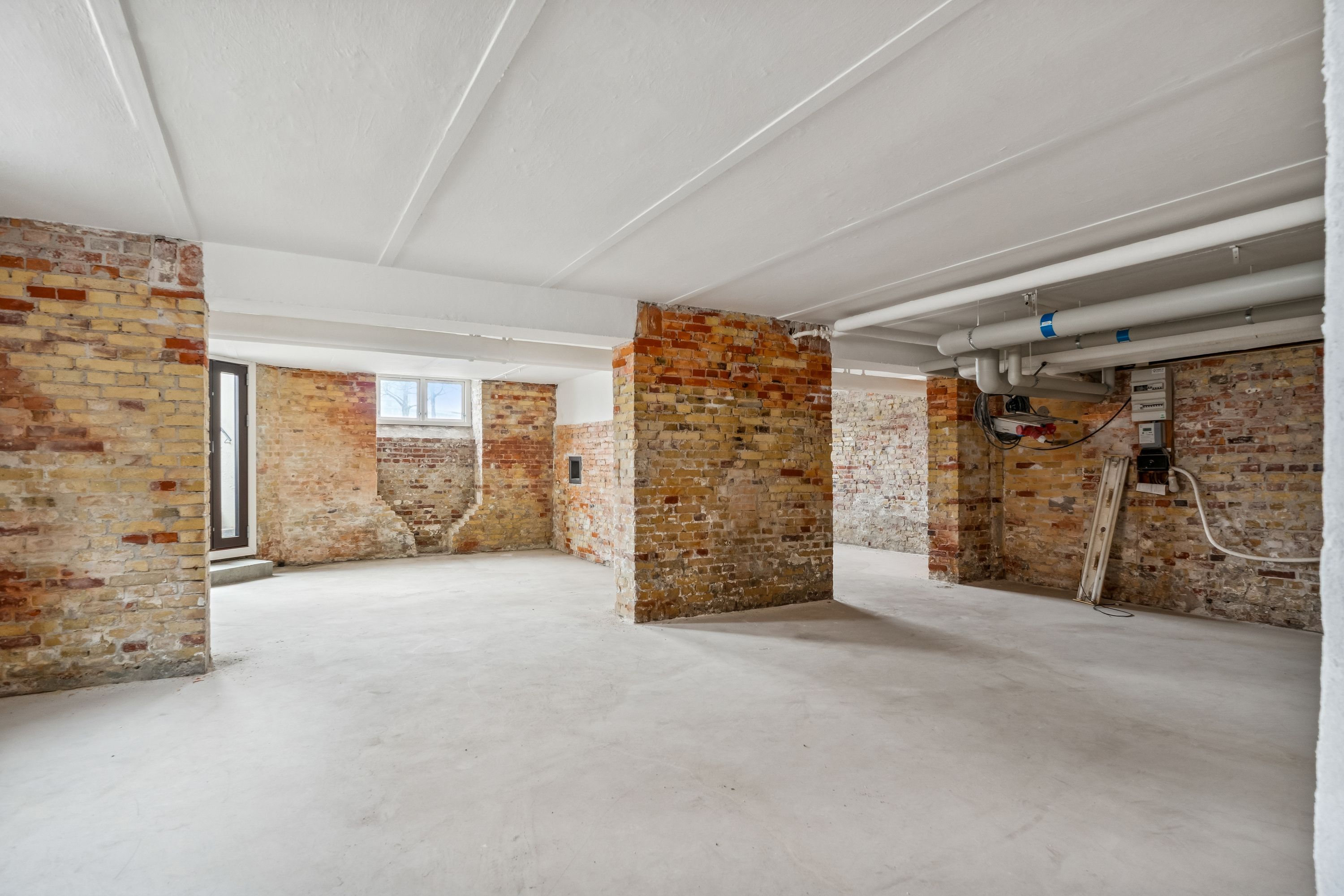 Lager til leje på Islands Brygge 19, København S. Lager med røde mursten og hvid loftsbeklædning. En stor vinduesåbning og tæt pakket rørledning.