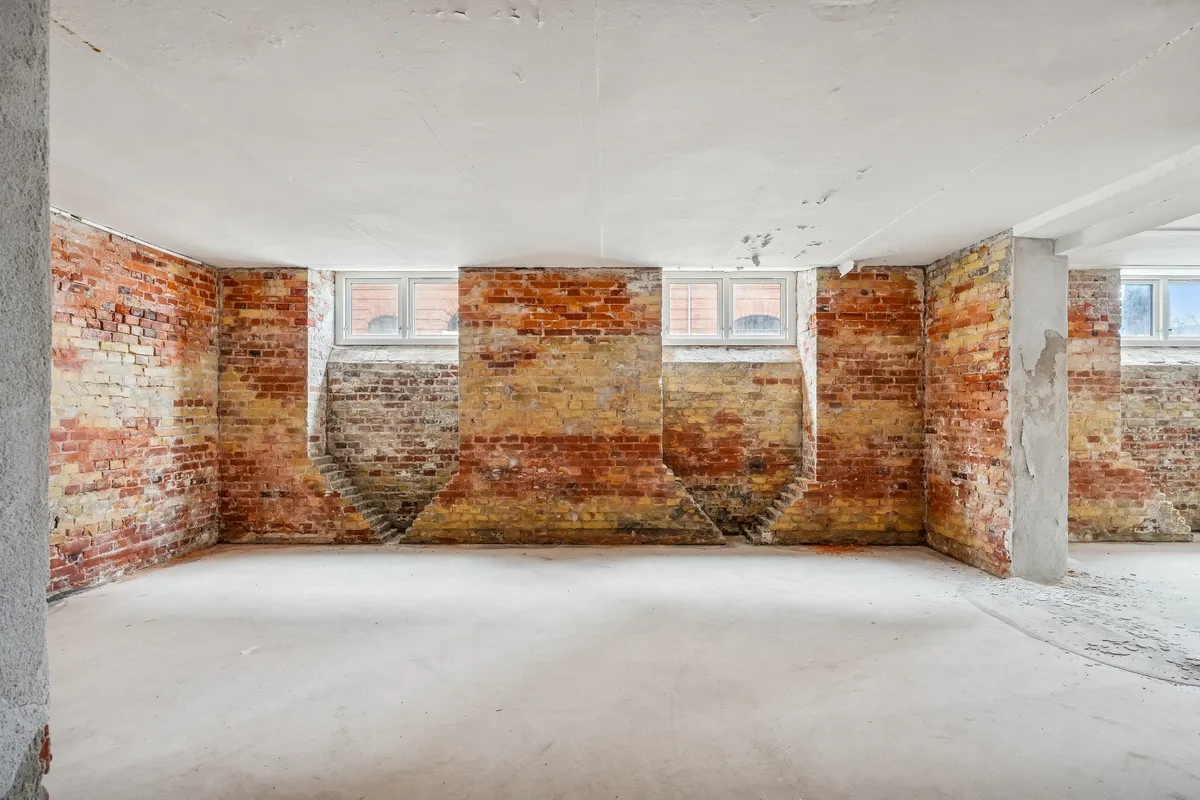 Lagerlokaler med rød mursten og hvid loftsbjælke. Hjørne med to vinduer og stige. Renset gulv og enkelte revner i murværket.