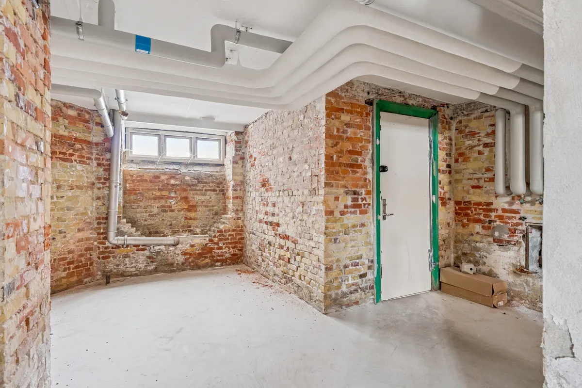 Lager med rød mursten og hvide loftsrør til leje på Islands Brygge 19. Rummet er tomt med grøn dør og rør i væggen.