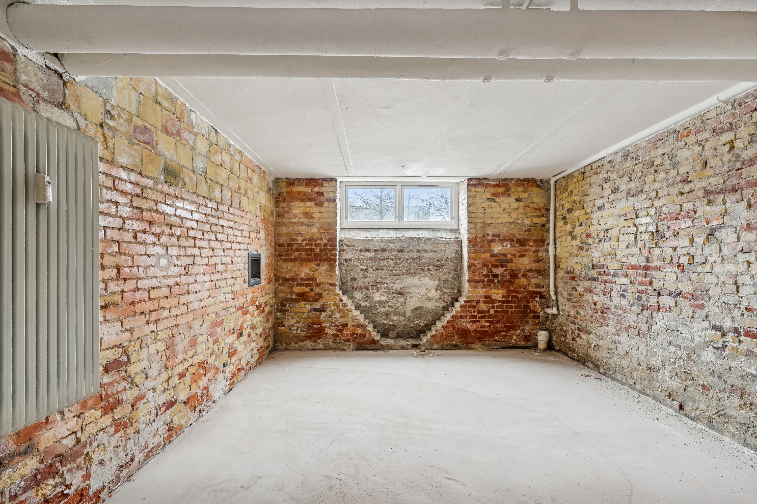 Lager til leje med røde murstensvægge, hvide loftsbjælker og gulv. Rum med en stor udvendig vindue og en indvendig trappe. Værelset er tomt og rent med et rødt gulv.