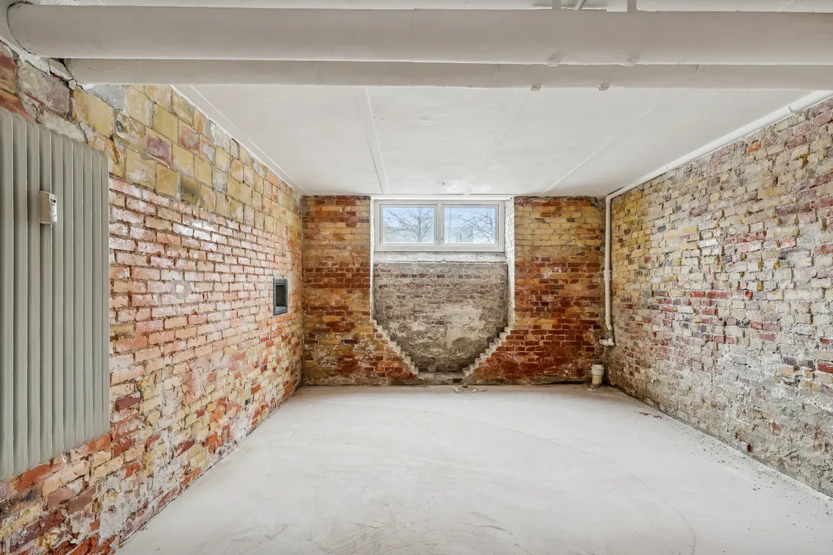 Lager til leje med røde murstensvægge, hvide loftsbjælker og gulv. Rum med en stor udvendig vindue og en indvendig trappe. Værelset er tomt og rent med et rødt gulv.