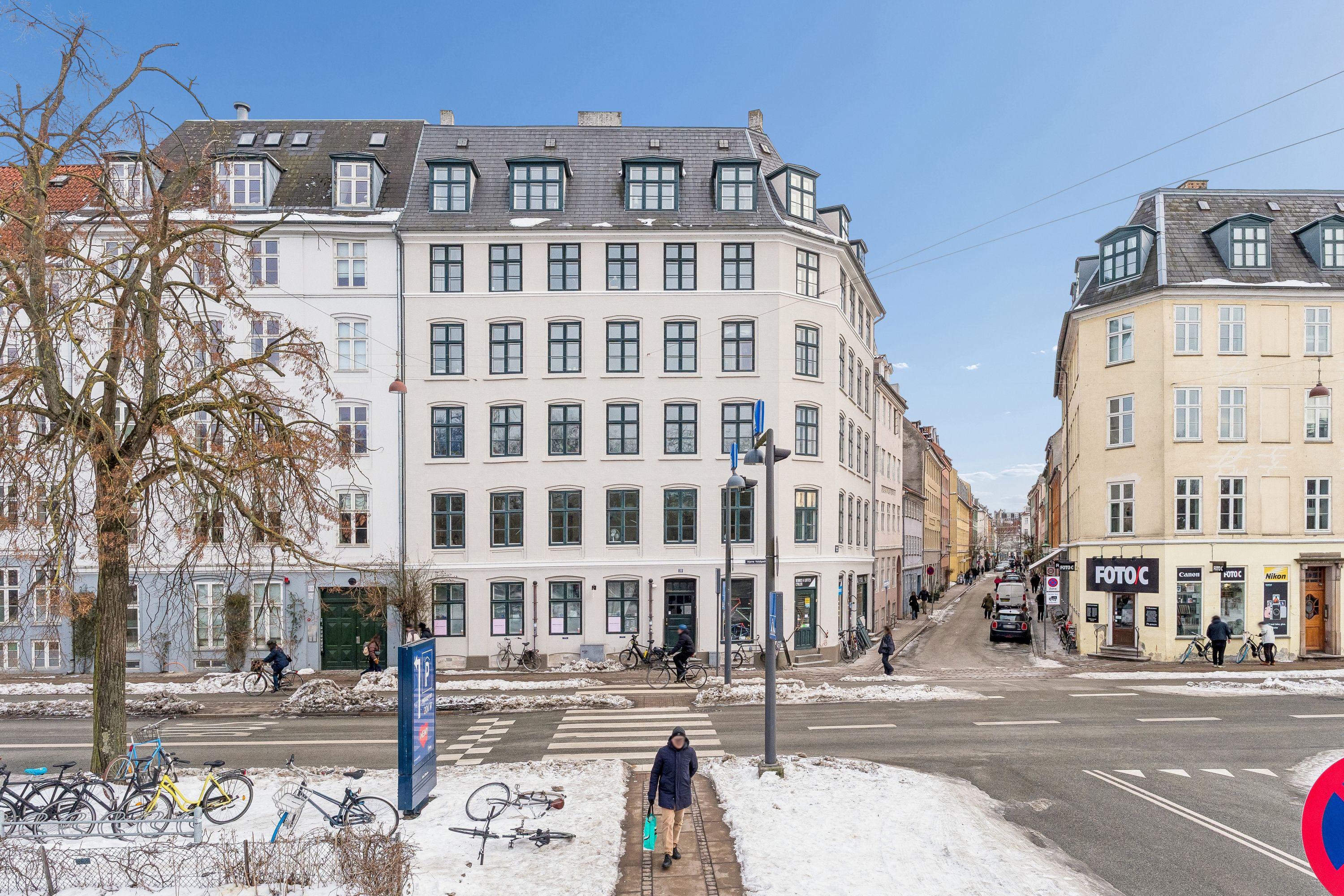 Butik til leje på Nørre Voldgade med adgang til parkeringsplads og cykelstel. Strømpebukser og sne på gaden og parkerede cykler. Folk går på fortovet og et foto- og videoforretningssted.