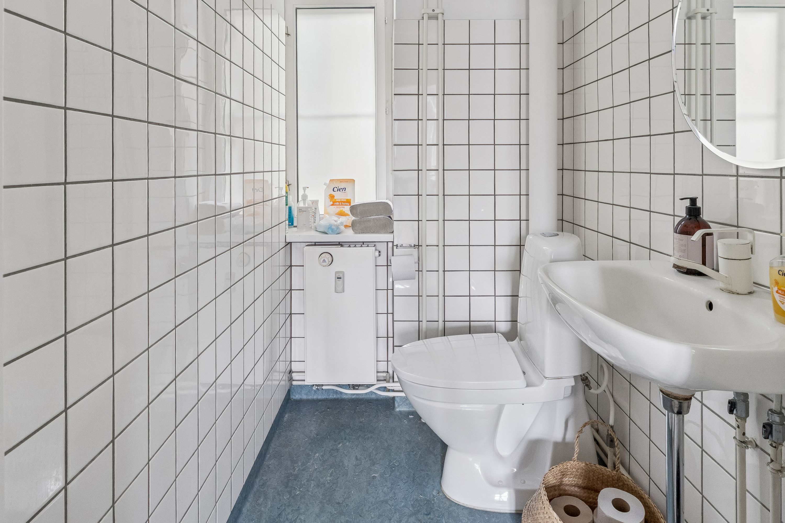 Toilette i hvidt badeværelse med spejl, håndvask, toilet, håndsæbe og to ruller toiletpapir i kurv