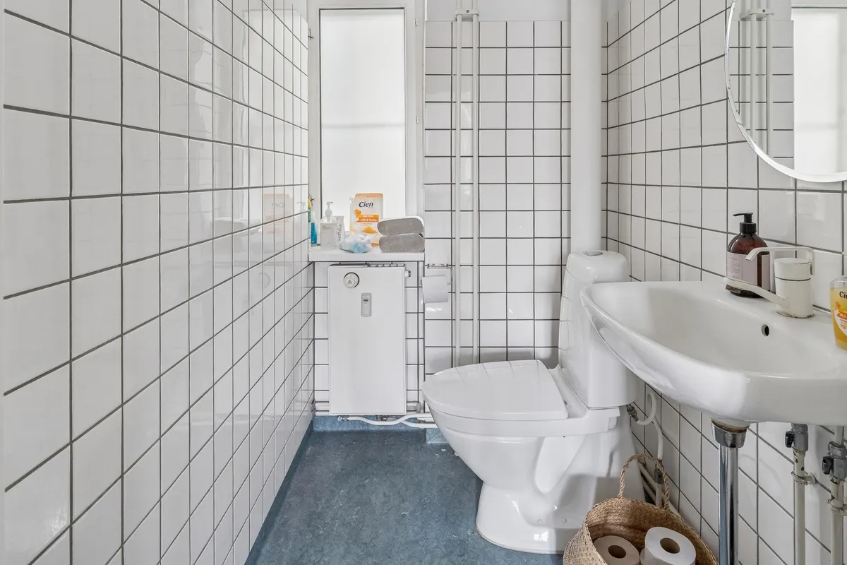 Toilette i hvidt badeværelse med spejl, håndvask, toilet, håndsæbe og to ruller toiletpapir i kurv