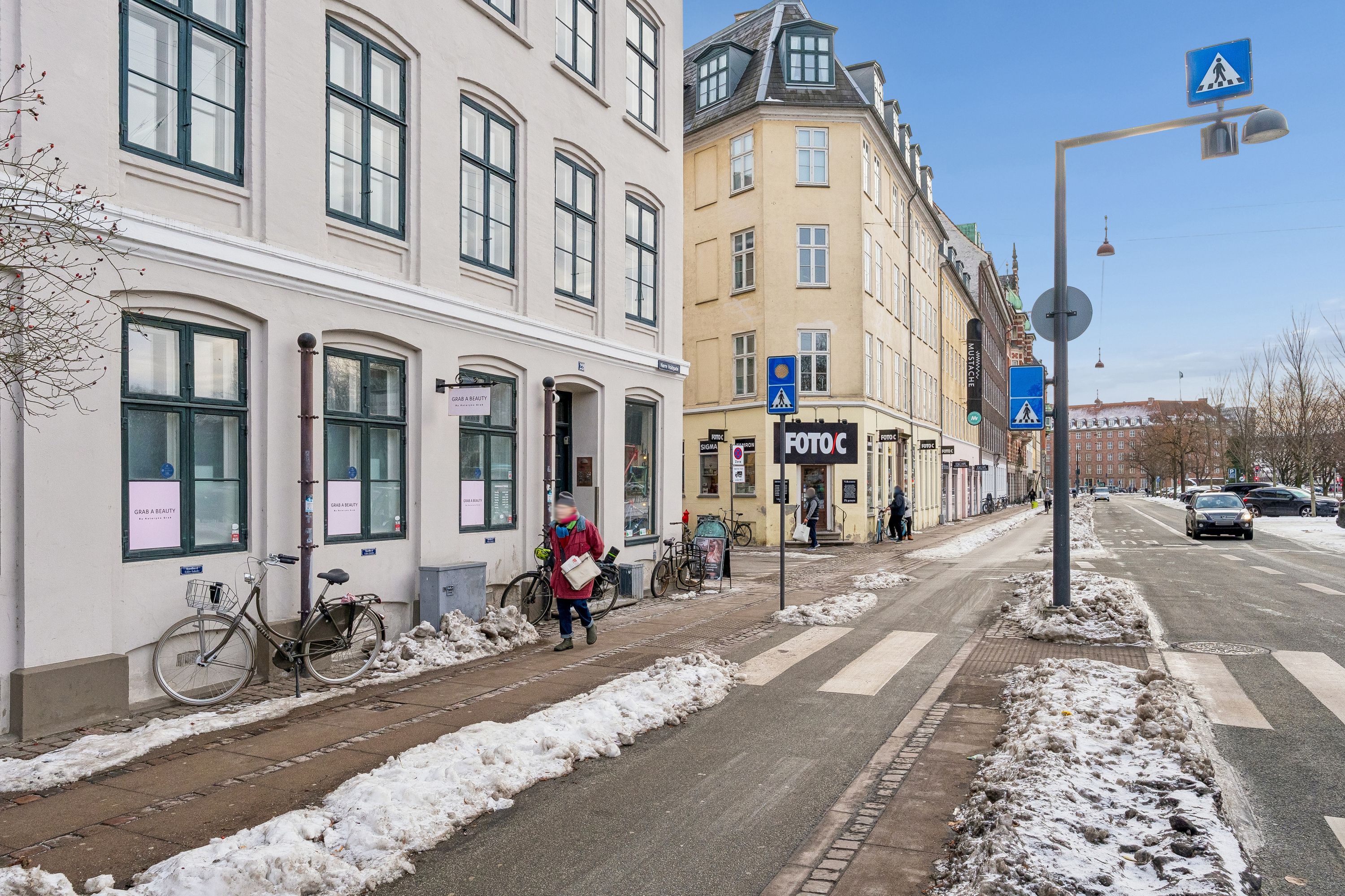 På snelagt gågade er der en person med hat og støvler. Butikkerne er forsynet med skilte, og der er flere cykler og biler. Søjlen med skilte og vejvisning står på hjørnet.
