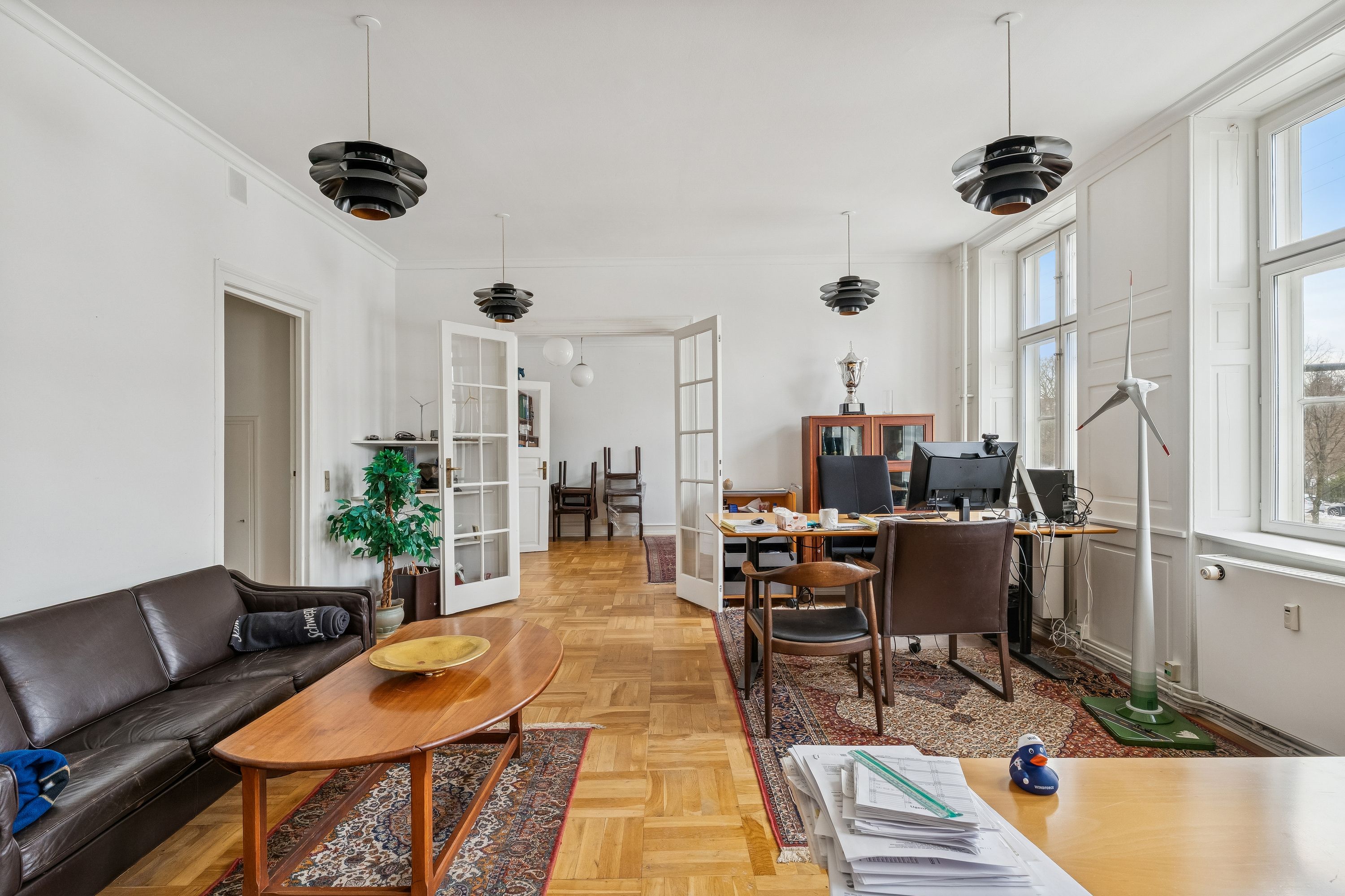 Lyst lokale med højt til loftet og højt gulv. To skriveborde med computere og møbler til kontorarbejde. Et lædersofa med et bord og en plante. To væghængte lamper og en vindmølle.
