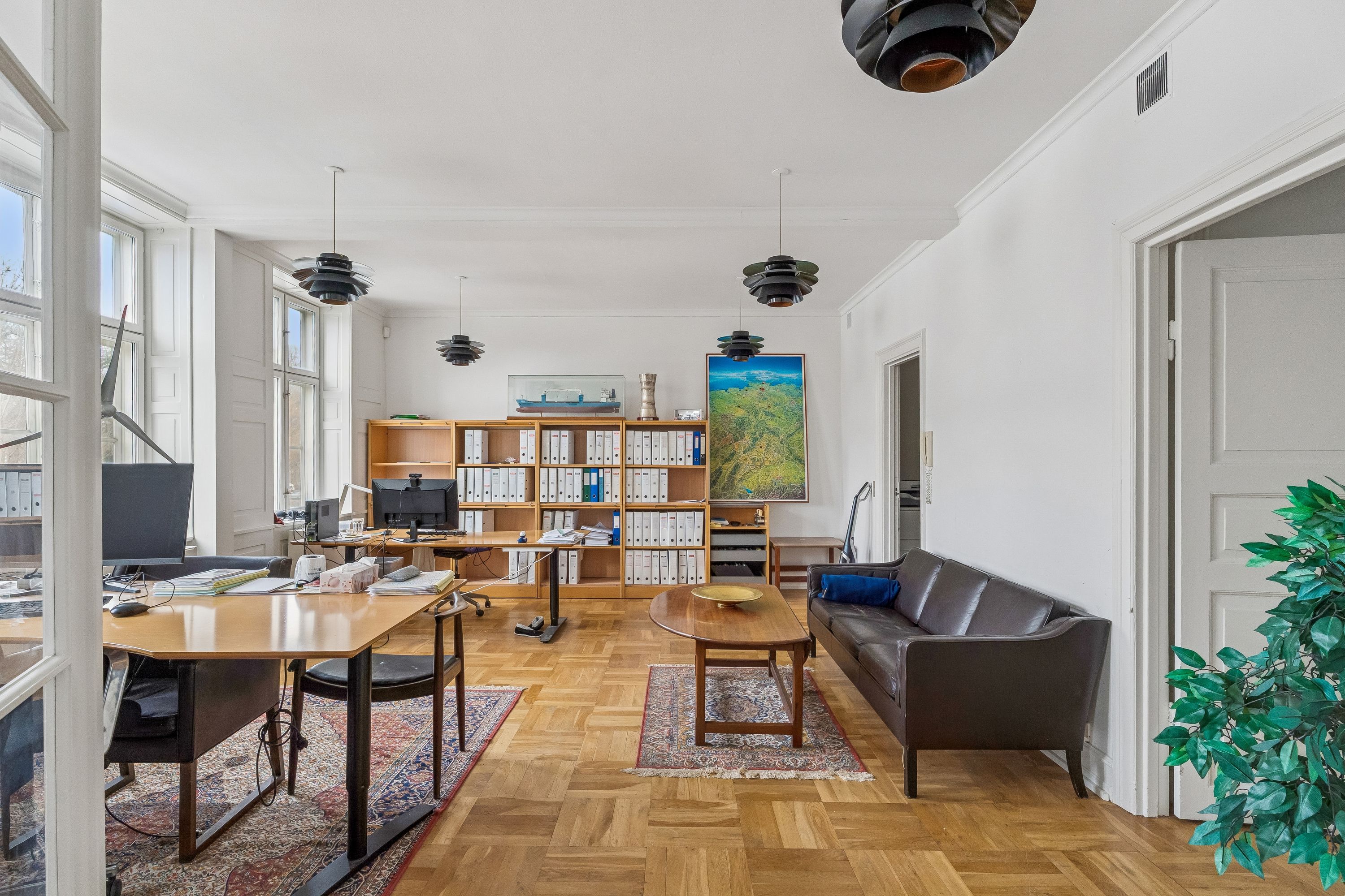 Kontor med skrivebord, stole, gulvlampe, sofa og kunstværk. Loft med moderne lysekroner og vinduer med udsigt til park og vej.