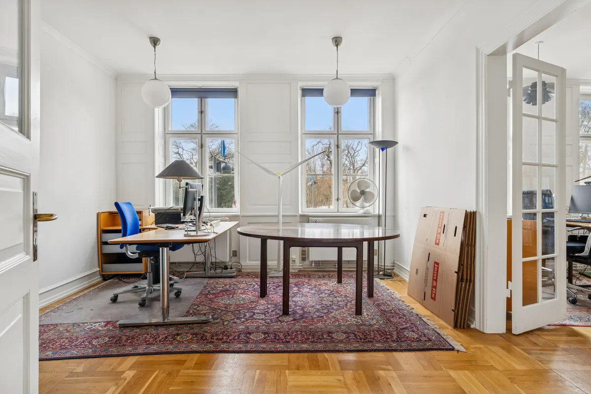 Et lille kontorlokal med moderne møbler og skriveborde, en stor orientalsk tæppe, hvide vægge, lyse gulve og slanke lamper samt en lyseblå stol