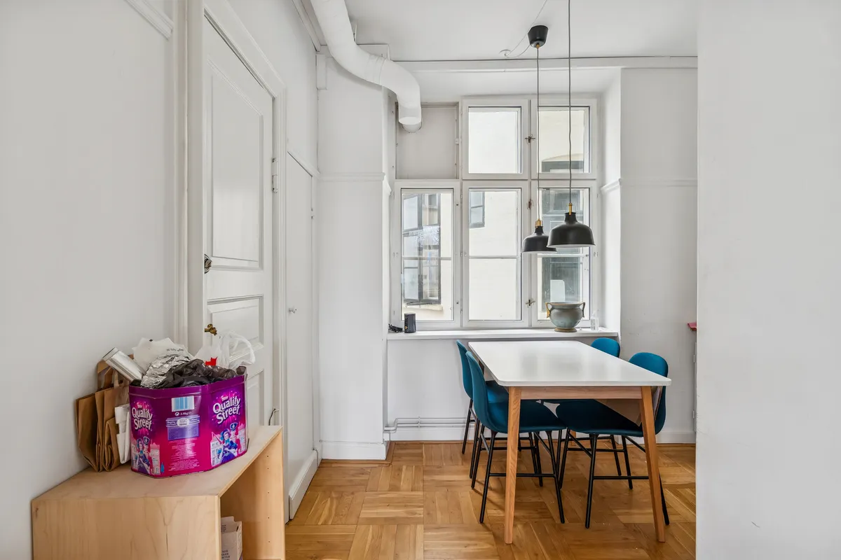 Alt: Lys stue med lysegult loft, hvid væg og trægulv. Bord med fire blå stole og lysekroner. Sort skraldespand og plastikposer på et bord.