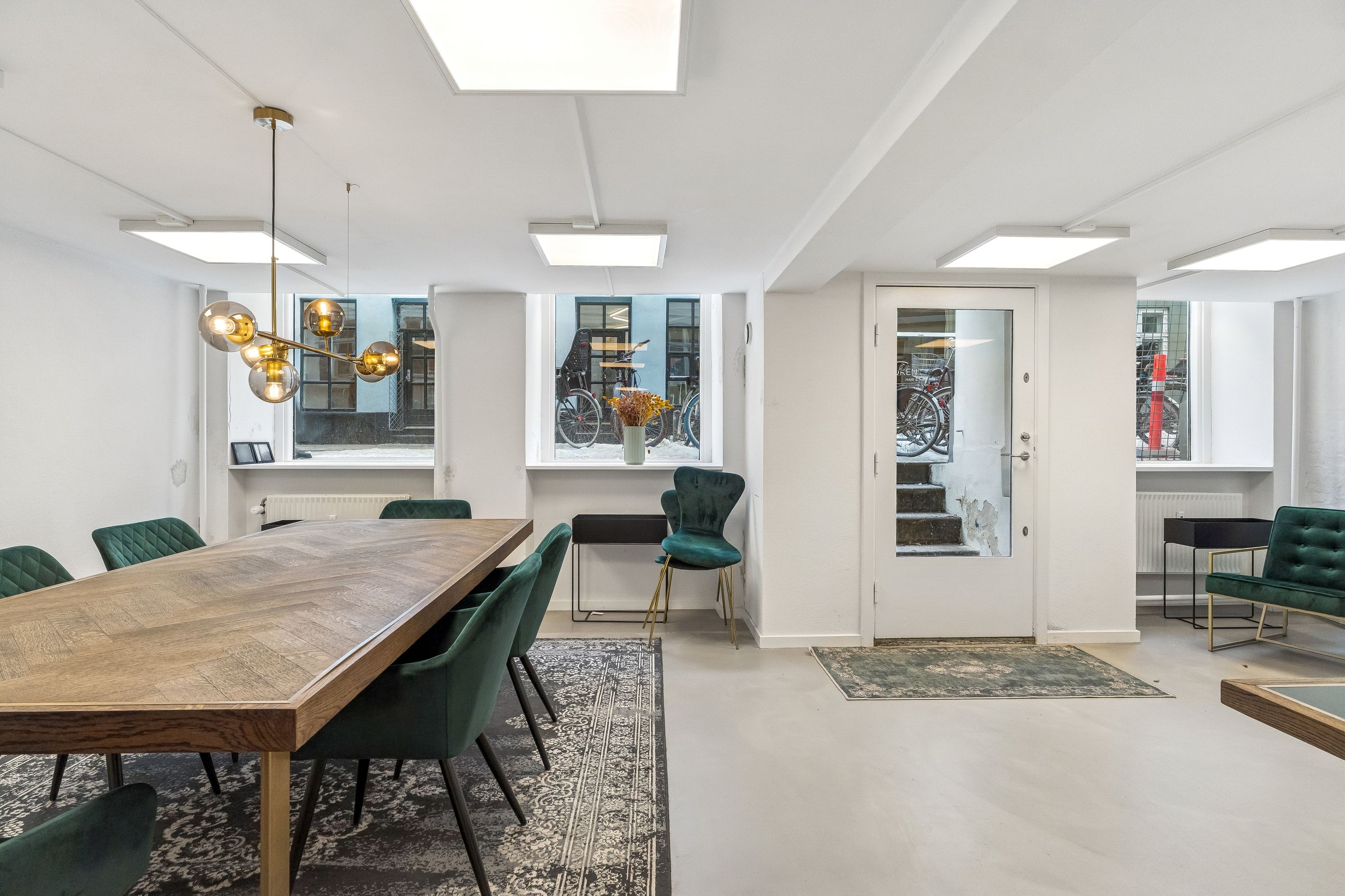 Kontor med langt træbord, gule gulvlampe og grønne stole samt 2 store vinduer med trappe og cykler udenfor