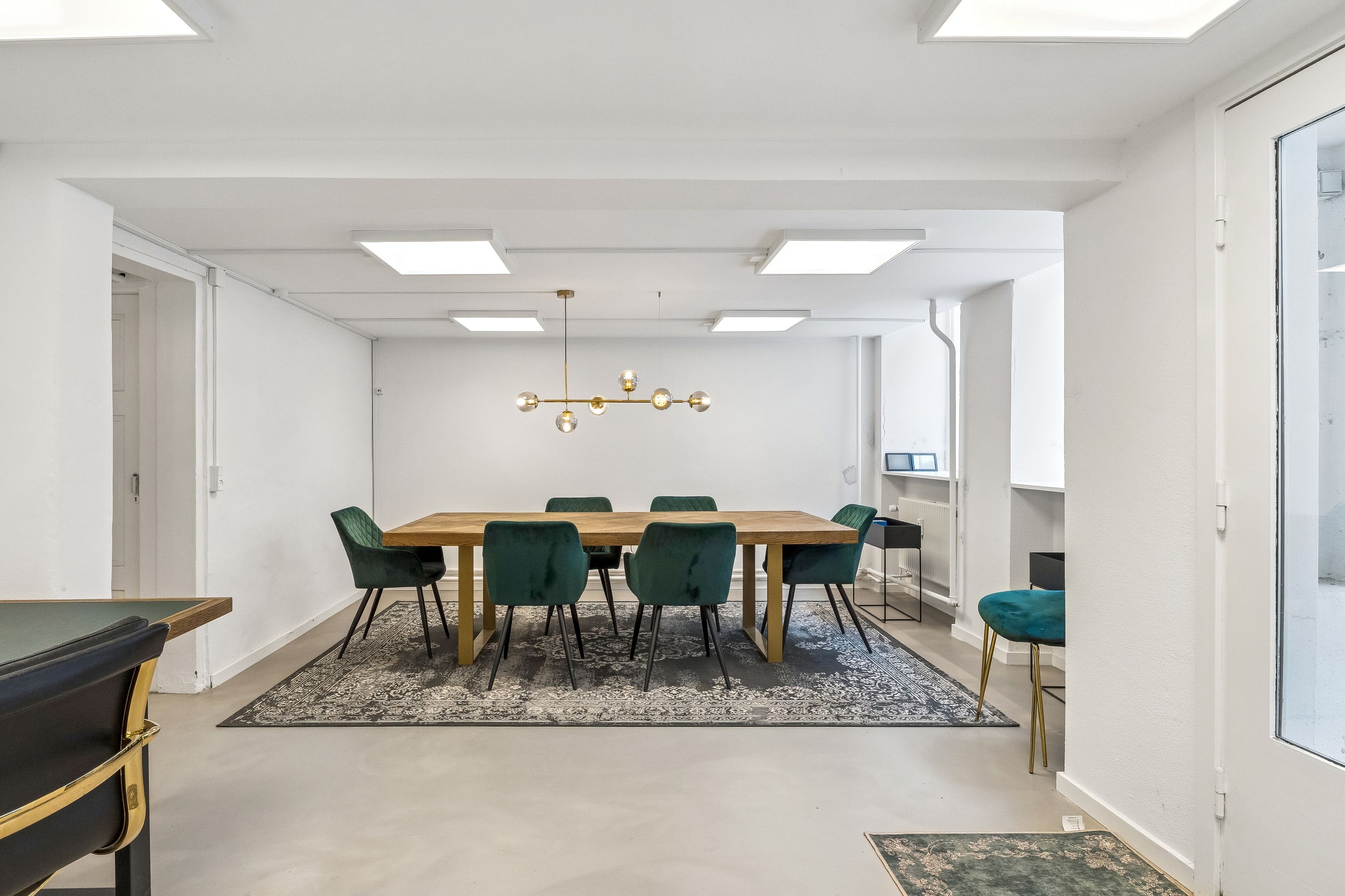 Lyst kontor med hvide vægge og lofter, et stort træbord med seks grønne stole, en elegant lysekron og et flot gulvtæppe i stuen