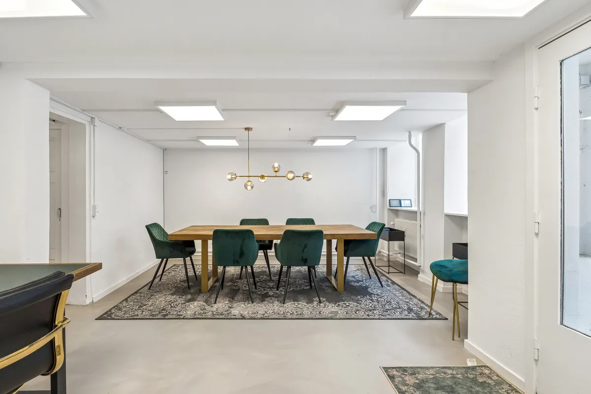 Lyst kontor med hvide vægge og lofter, et stort træbord med seks grønne stole, en elegant lysekron og et flot gulvtæppe i stuen