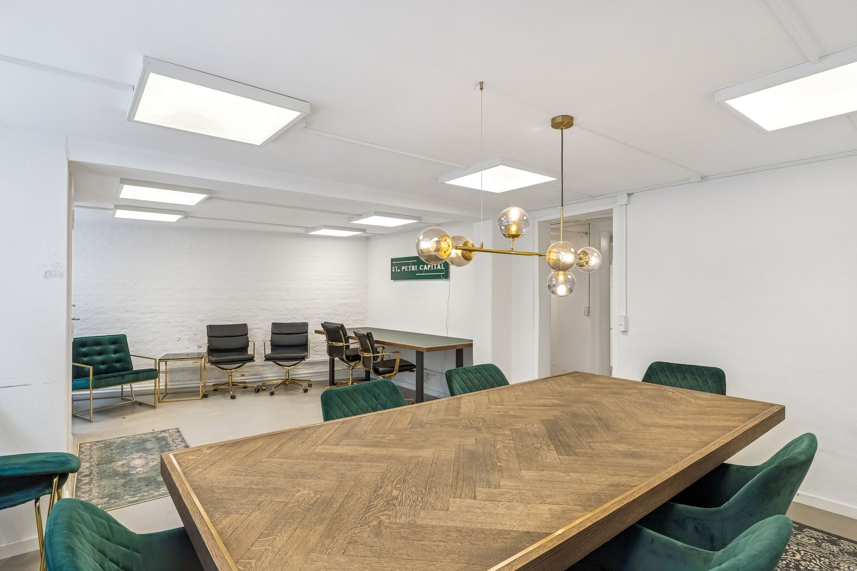 Moderne mødelokale med lyse lofter, et stort bord og fire grønne stole med guldbeslag samt et lysekron med runde lamper