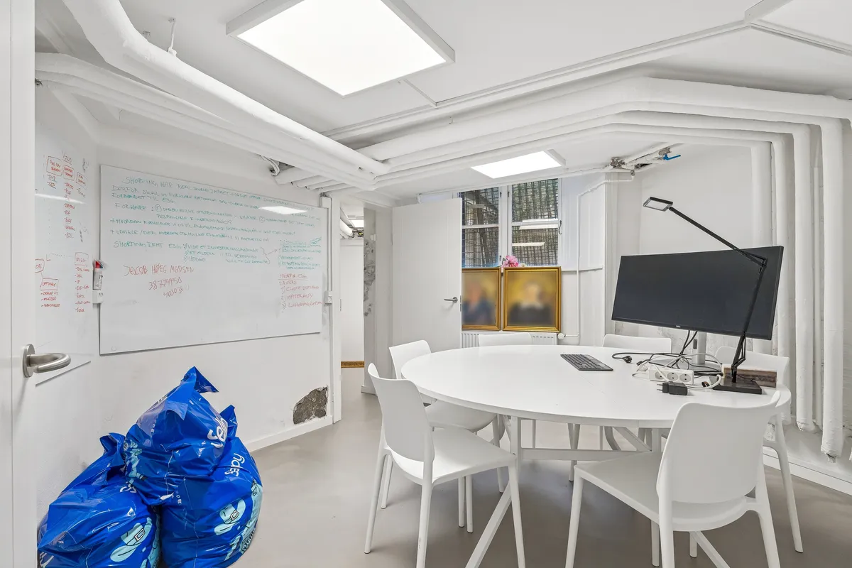Et kontorruum med hvide vægge, gulve og lofter, hvor der er en rund hvid bord med flere stole og en computer med en monitor og lampe. Der er en hvid tavle med tekst og billeder.
