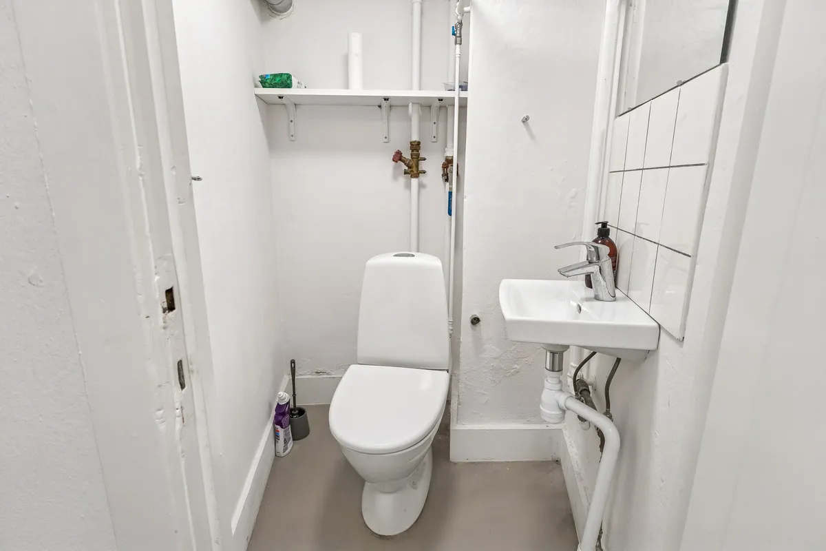 En lille toiletrum med hvide vægge og lofter og en hvid toilet, hvor der er en håndvask og en håndsæbeflaske på væggen
