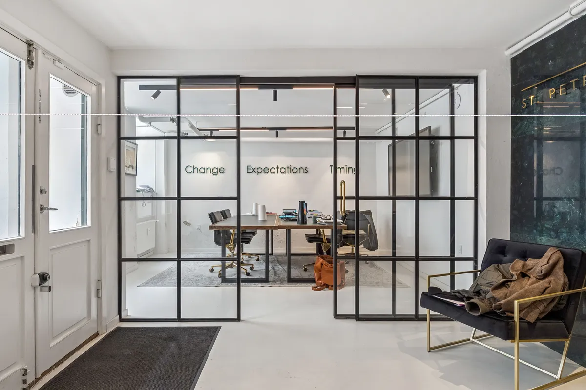 En hvid indgangsdør med sølvbeslag, en bordsætning med to borde og fire stolpe, samt en gulvlampe i guld. Et sort og guldt farvet stol med en beige jakke på ryggen