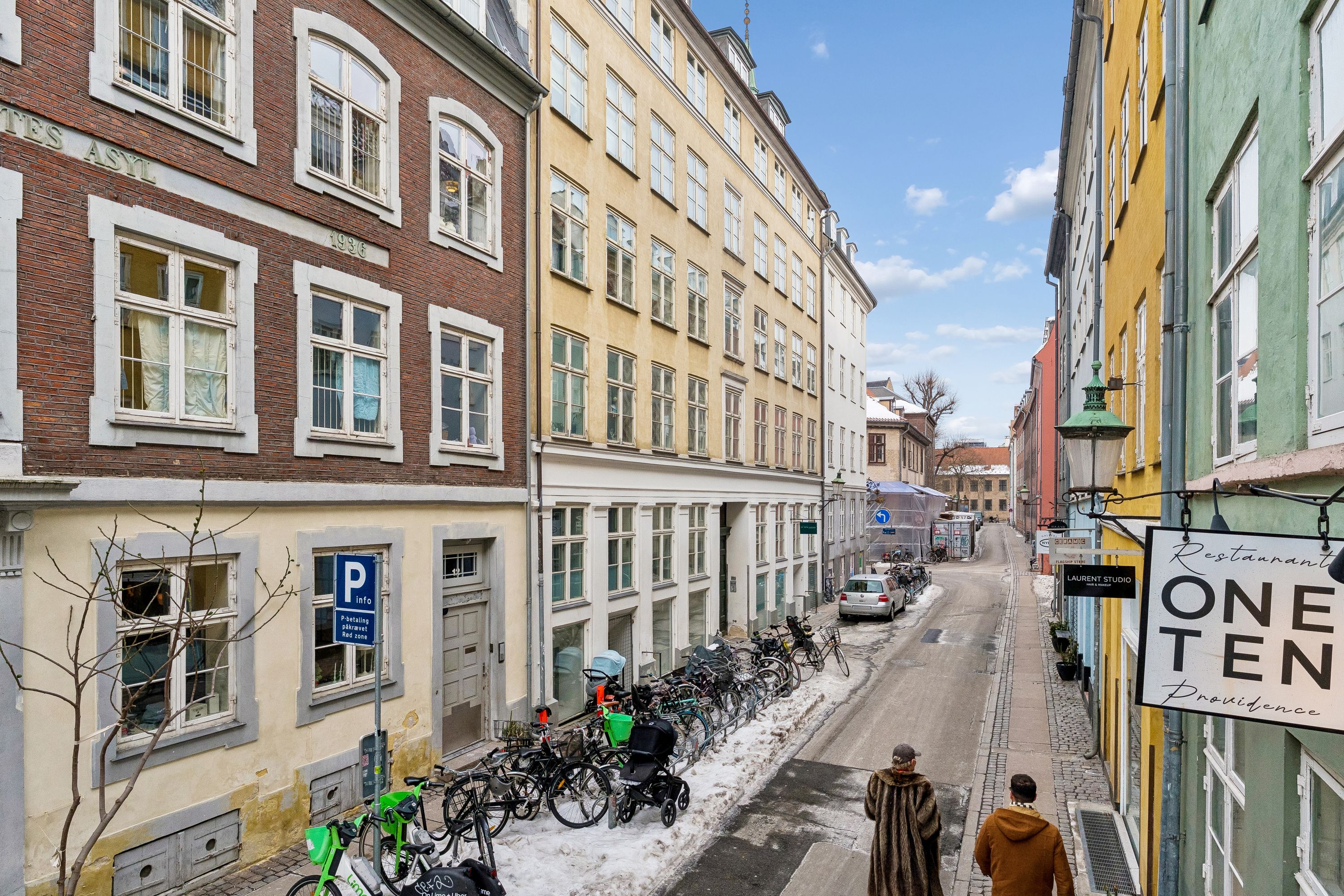 To mennesker går ned ad en smal gade med byggeskilte og parkerede cykler. På højre side er en butik med skiltet "ONE TEN Providence" og en grøn lampe. Baggrund: blå himmel med skyer og træer.