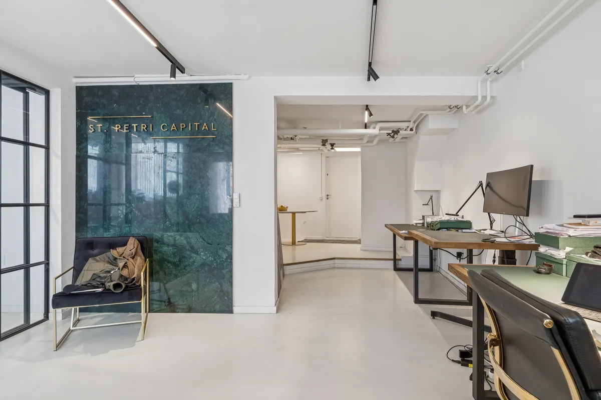 Kontorlokaler med minimalistisk interiør. Marmorvæg med logoet St. Petri Capital. Kontorbord med computer, lampe og papirkurve. Lædermøbel med guldkant. Væggen er hvid med glasruder og lysekroner.